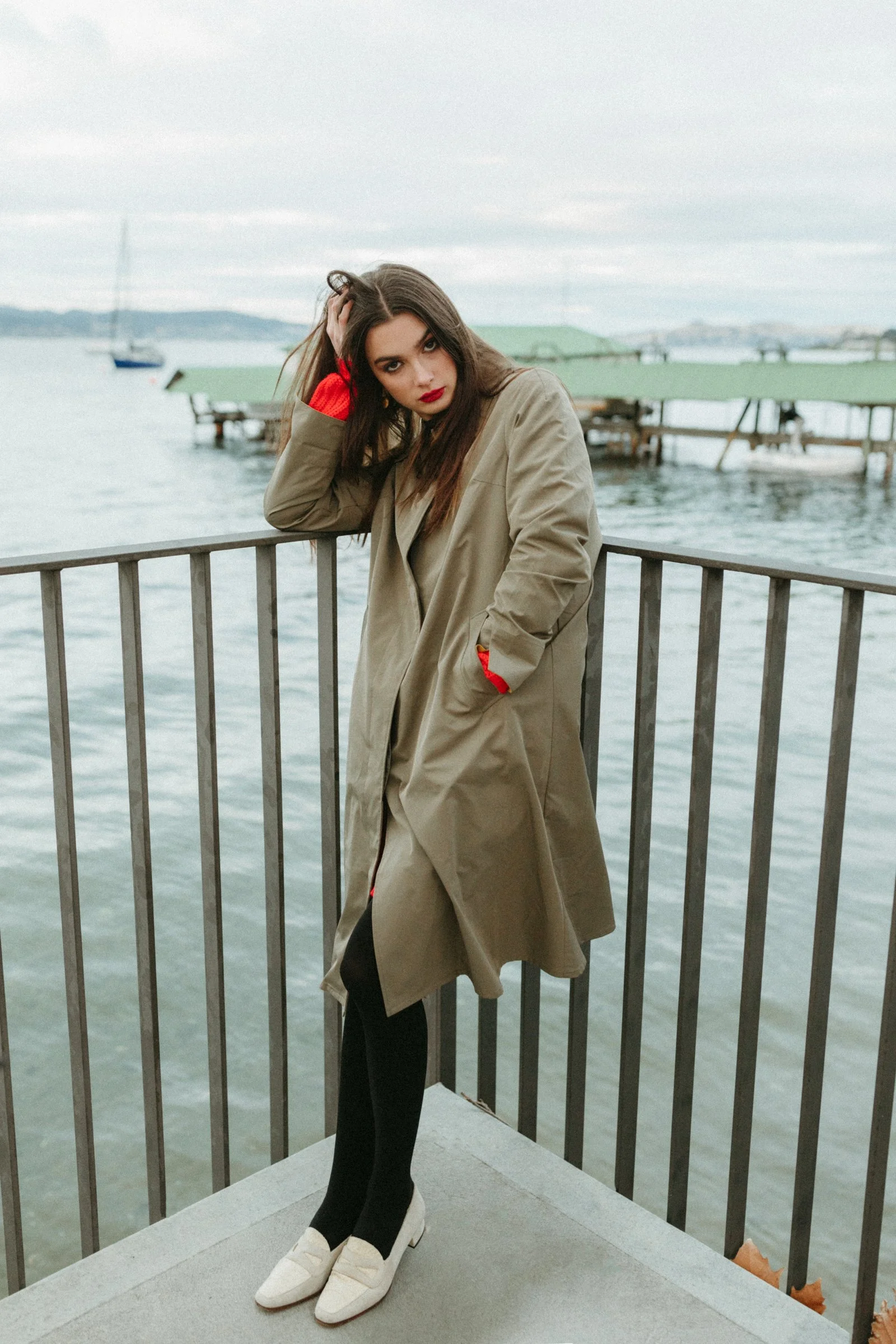 Model poses by ocean in trench coat.