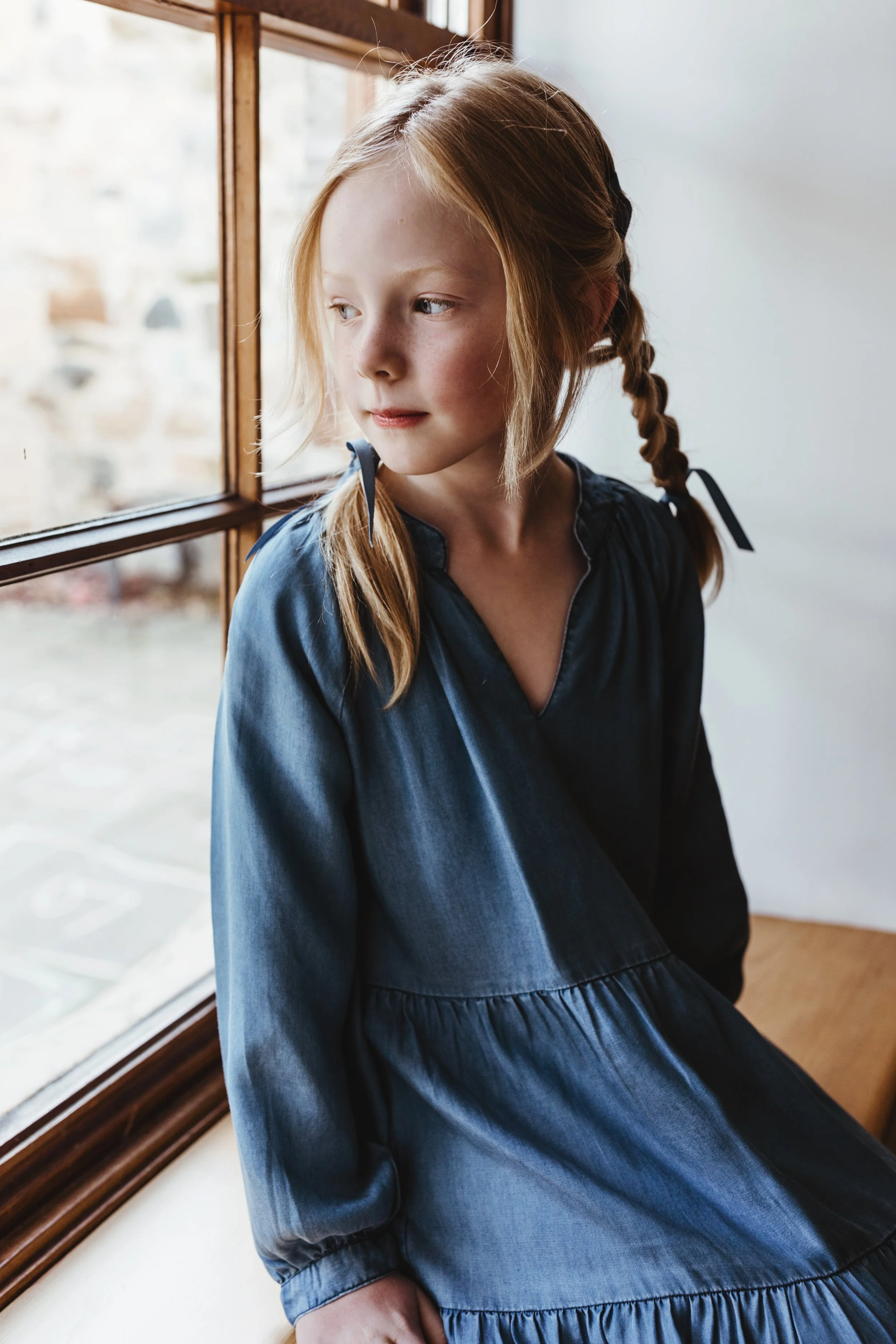 Young girl looks out of window in denim dress for commercial photo shoot.
