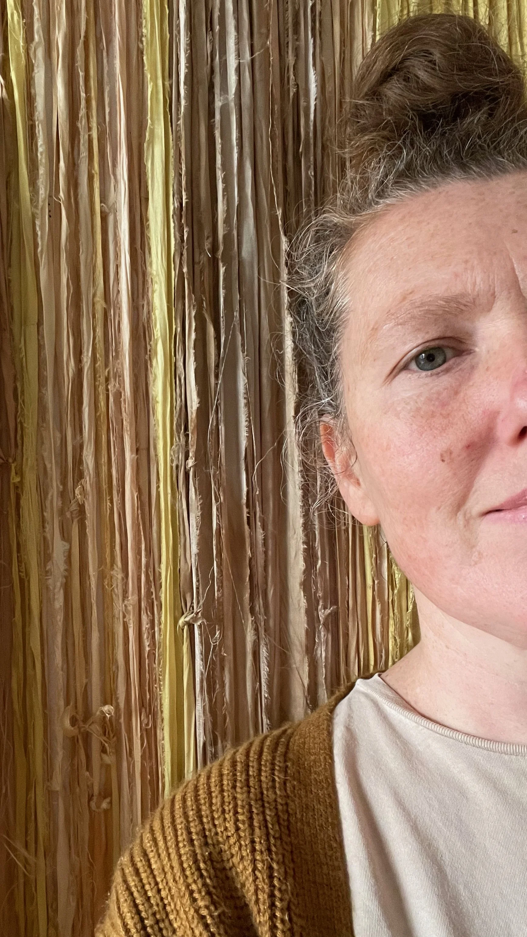 Close-up of a woman's face on the right side of the image, with a textured brown cardigan, against a background of a wall made of intertwined natural ropes or twine.