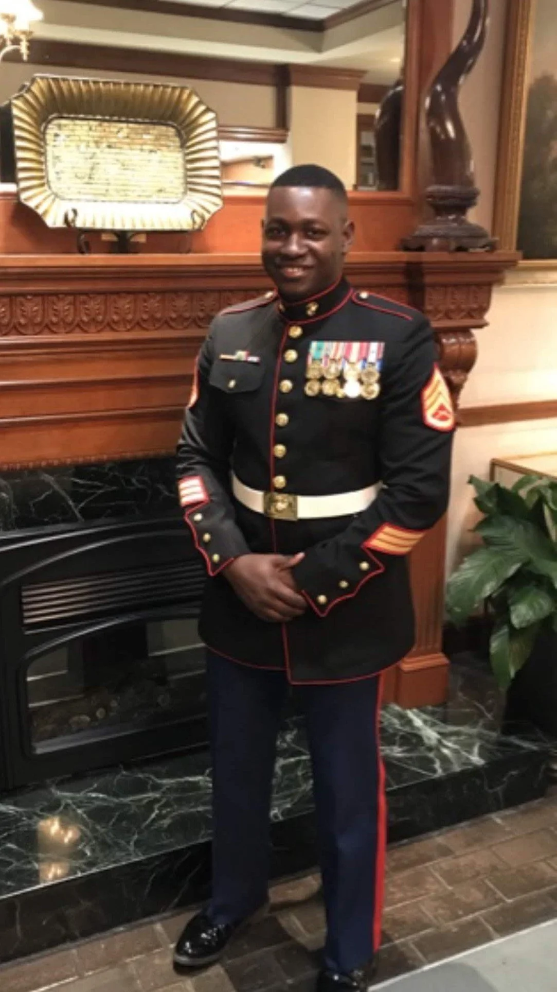 A man in a formal military uniform with medals and insignia, standing indoors near a fireplace and smiling.