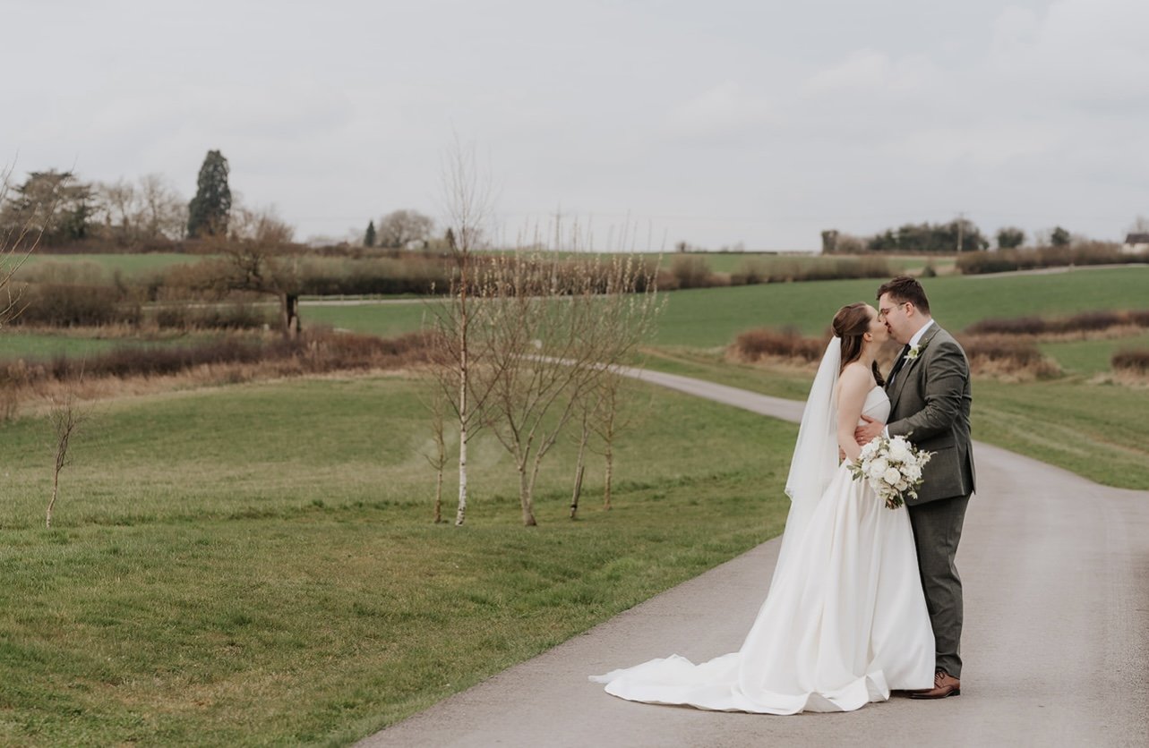 A bride and groom sharing a kiss outdoors on a winding road, with green fields and trees in the background.