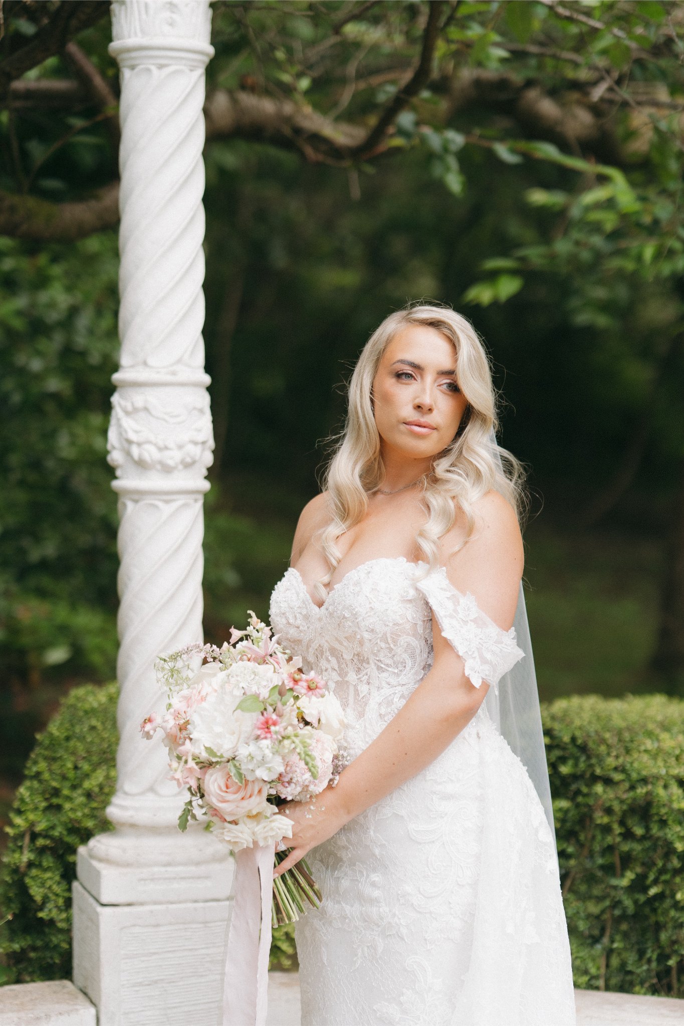 A bride in a white lace wedding gown holding a pink and white bouquet, standing outdoors next to a white twisted column with greenery in the background.