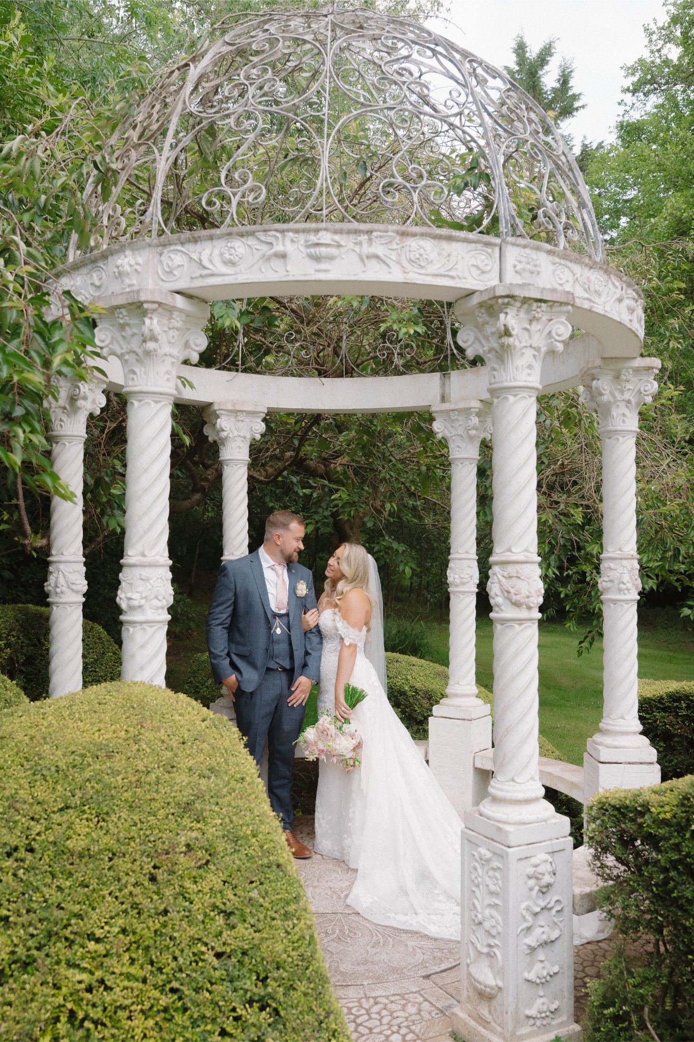 A bride and groom stand together under a white ornate gazebo in a garden, sharing a moment on their wedding day. The bride is wearing a white wedding dress and holding a bouquet, while the groom is dressed in a blue suit with a pink tie.