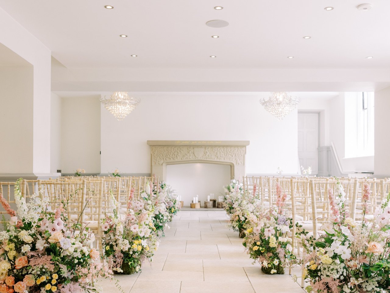 Elegant wedding ceremony setup with rows of light-colored chairs, floral arrangements, a fireplace, and chandeliers in a bright, white room.