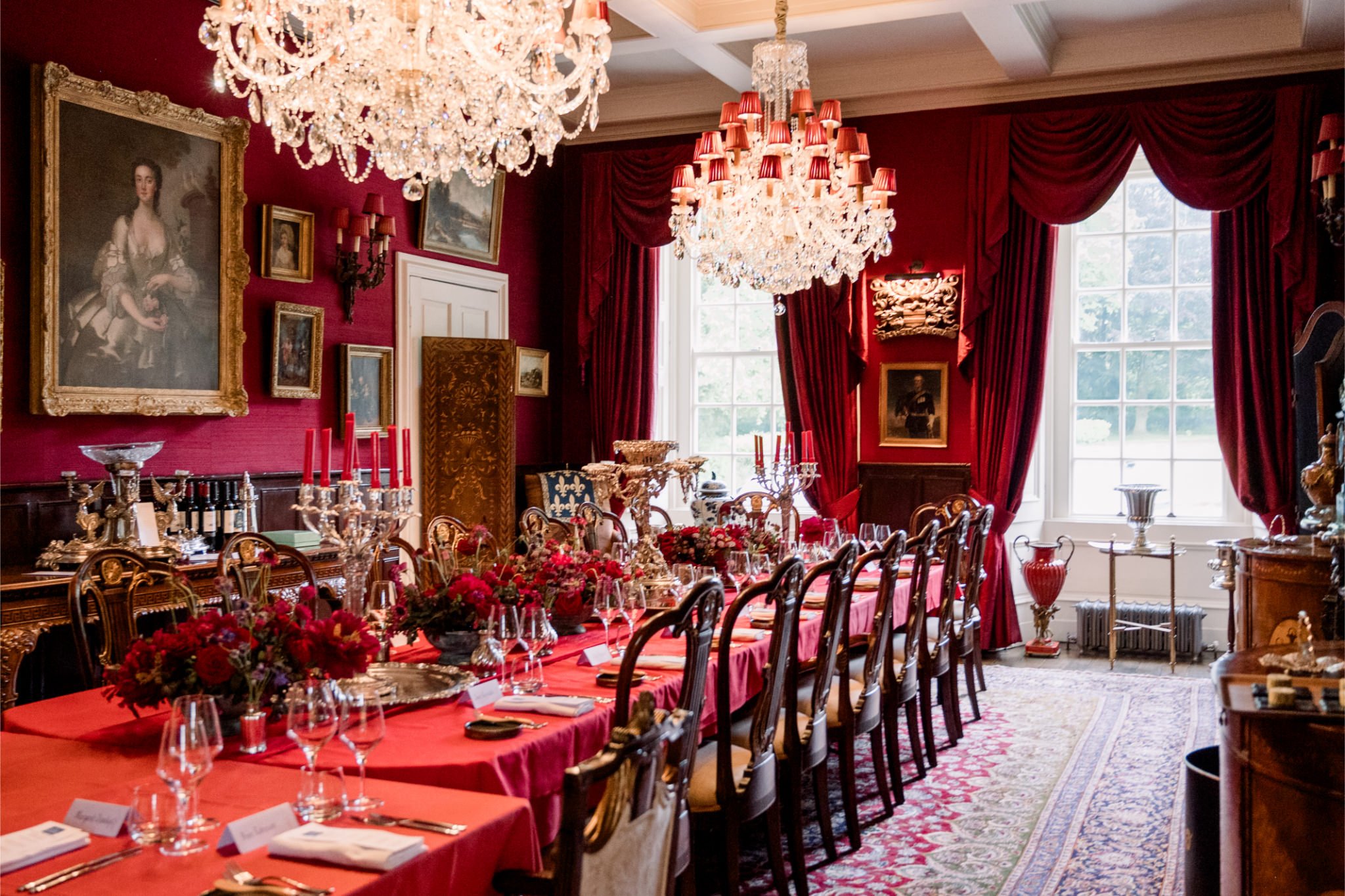 Elegant dining room with a long red tablecloth-covered table, ornate wood chairs, floral centerpieces, crystal chandeliers, historical paintings on red walls, large windows with red curtains, and decorative accents.