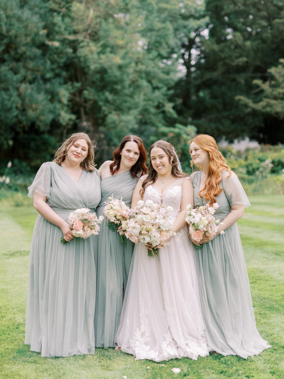 Four women dressed in elegant dresses standing outdoors on a green lawn, holding bouquets of flowers with trees in the background. One woman is wearing a white wedding gown, indicating a wedding celebration.
