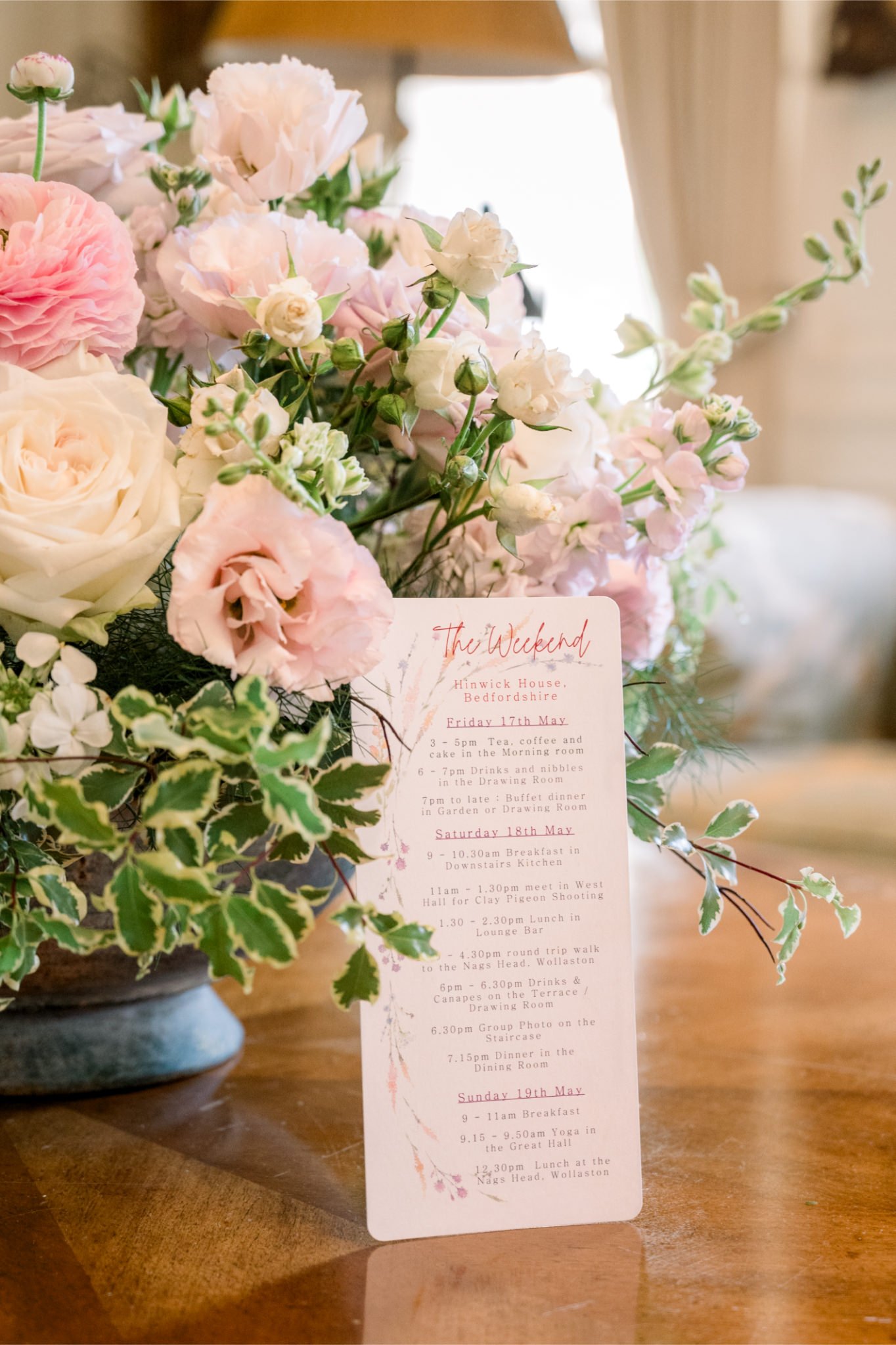 A floral arrangement with pink and white flowers, including roses and lisianthus, alongside a schedule card with a weekend itinerary, placed on a wooden table in a cozy indoor setting.