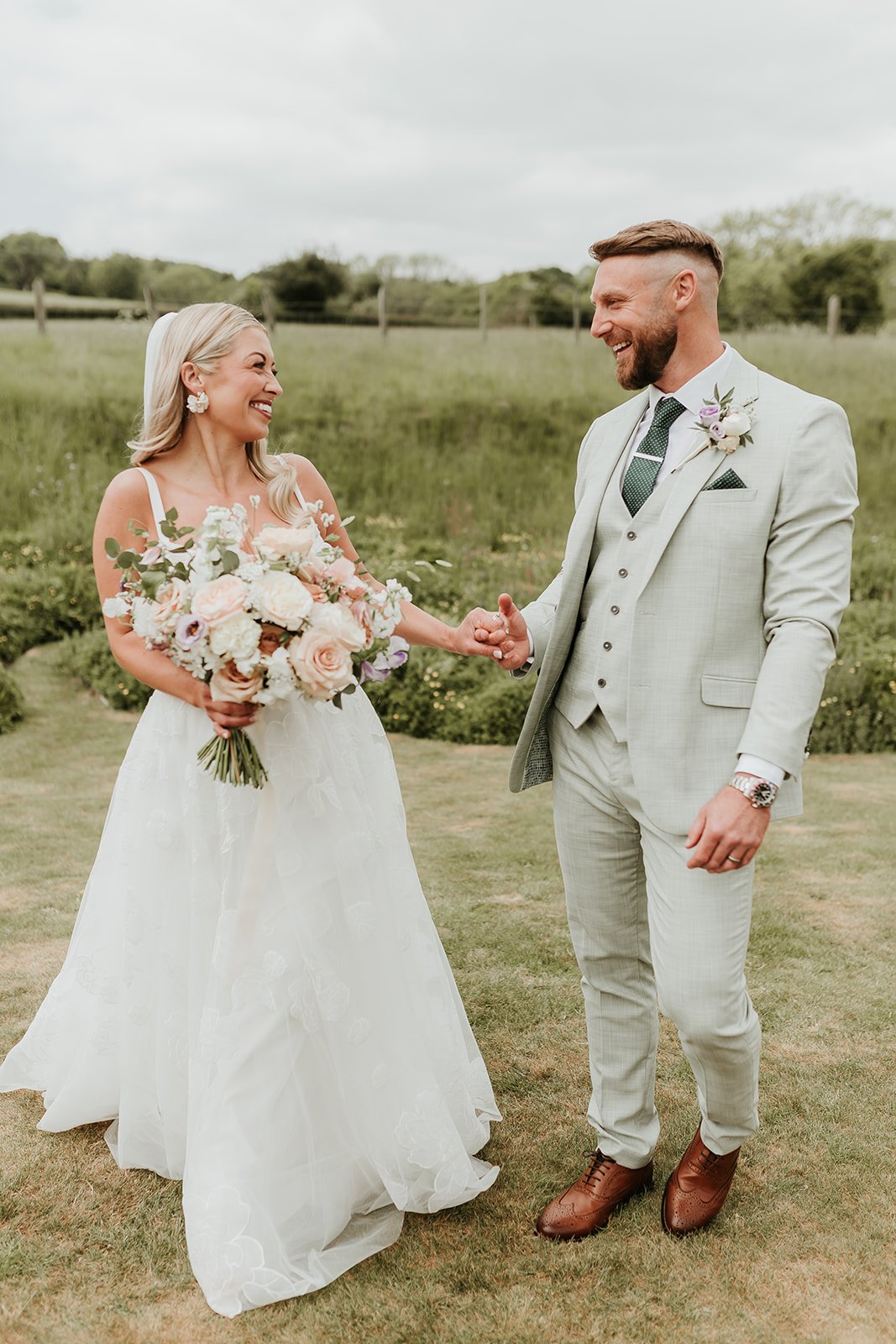Bride and groom holding hands and smiling outdoors during their wedding ceremony, green field background.