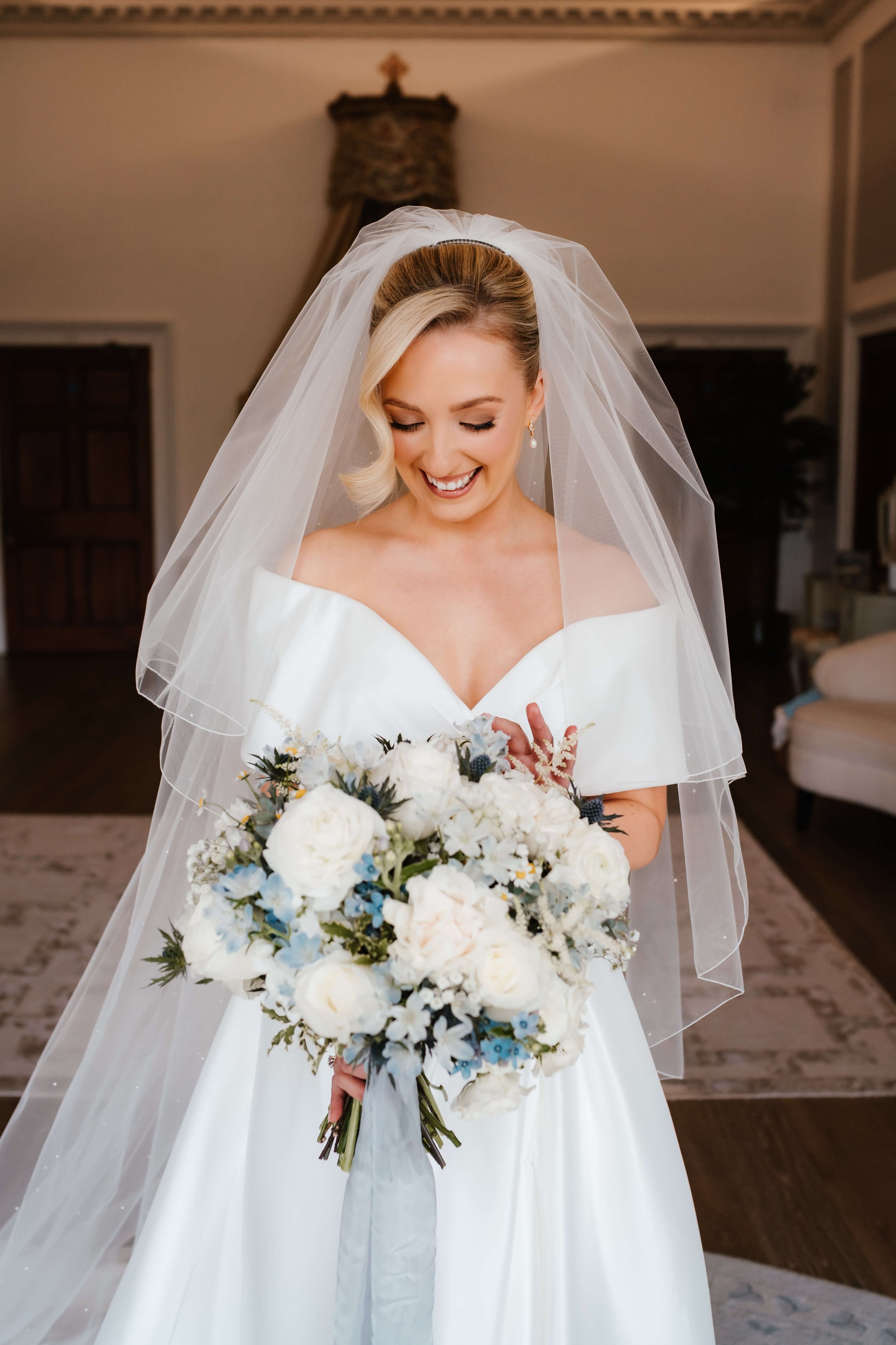 A smiling bride wearing an off-the-shoulder white wedding gown and veil, holding a large bouquet of white and light blue flowers, standing indoors in a warmly lit room.
