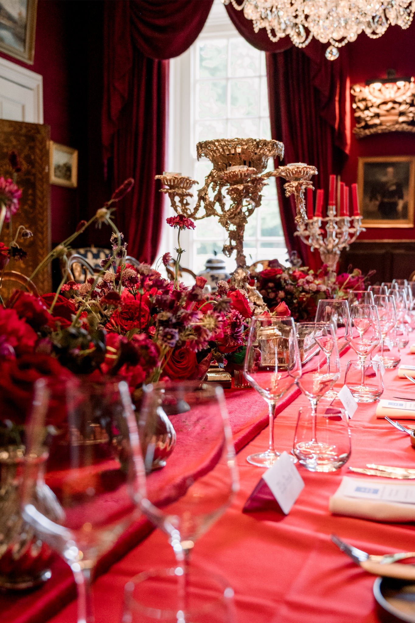 A luxurious dining table decorated with red flowers, crystal candelabras, and tableware, set in a room with deep red walls, gold accents, and a large window with heavy red curtains.