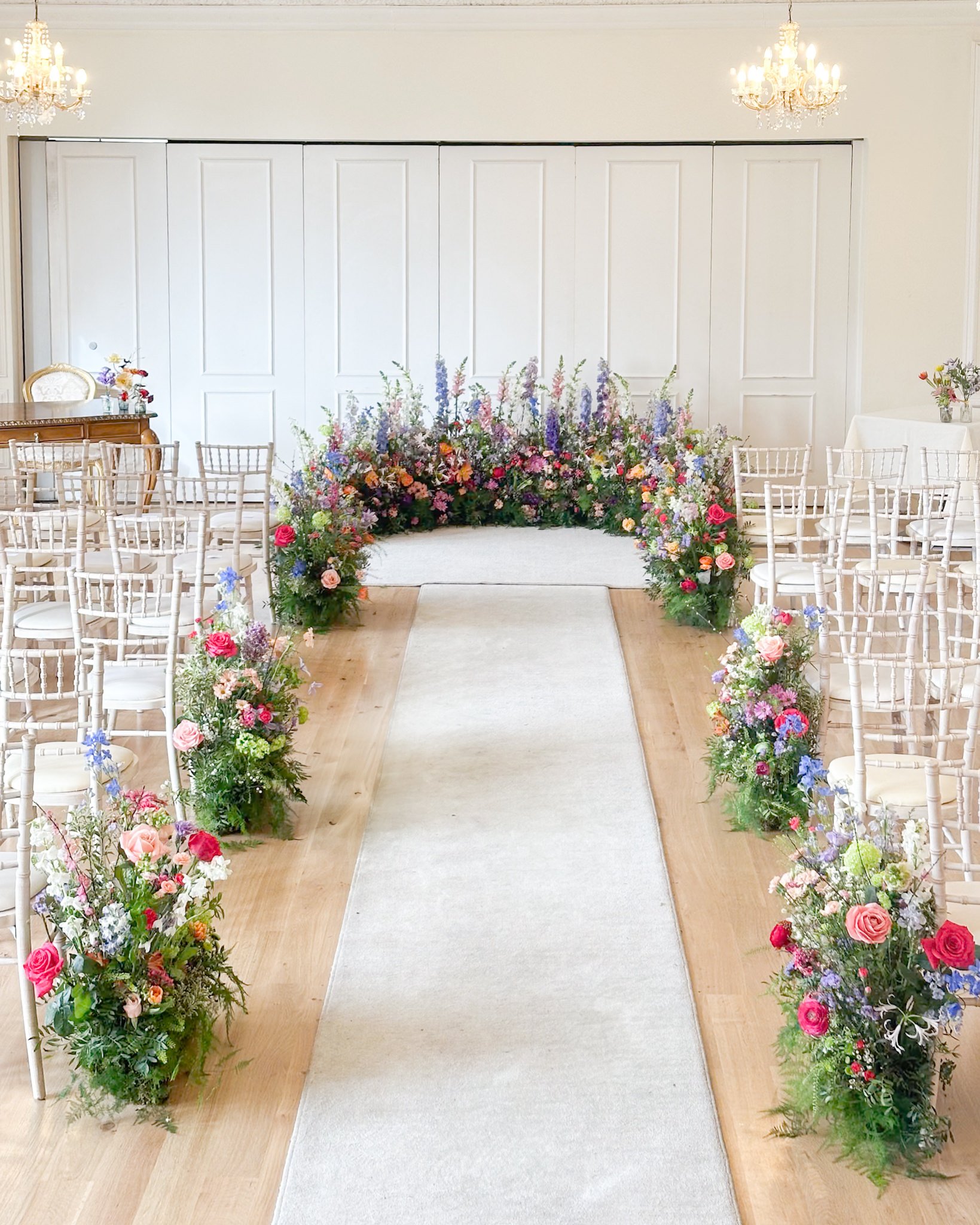 A wedding ceremony aisle decorated with colorful floral arrangements on both sides, leading to a rose flower arch at the front, in an elegant room with chandeliers and white walls.