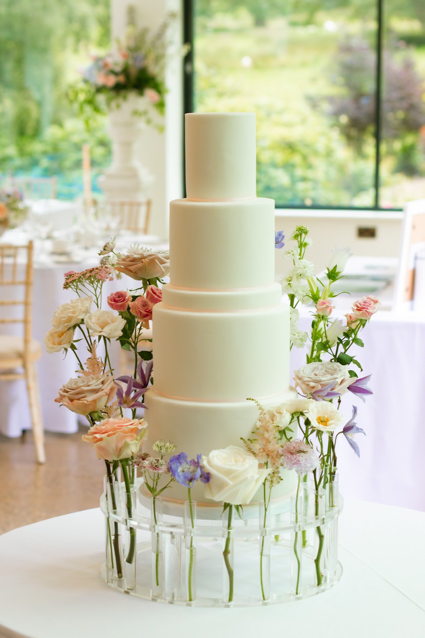 A four-tier white wedding cake with pastel pink accents, decorated with various white and light pink flowers, surrounded by a clear glass flower holder with additional flowers at the base on a round white table.