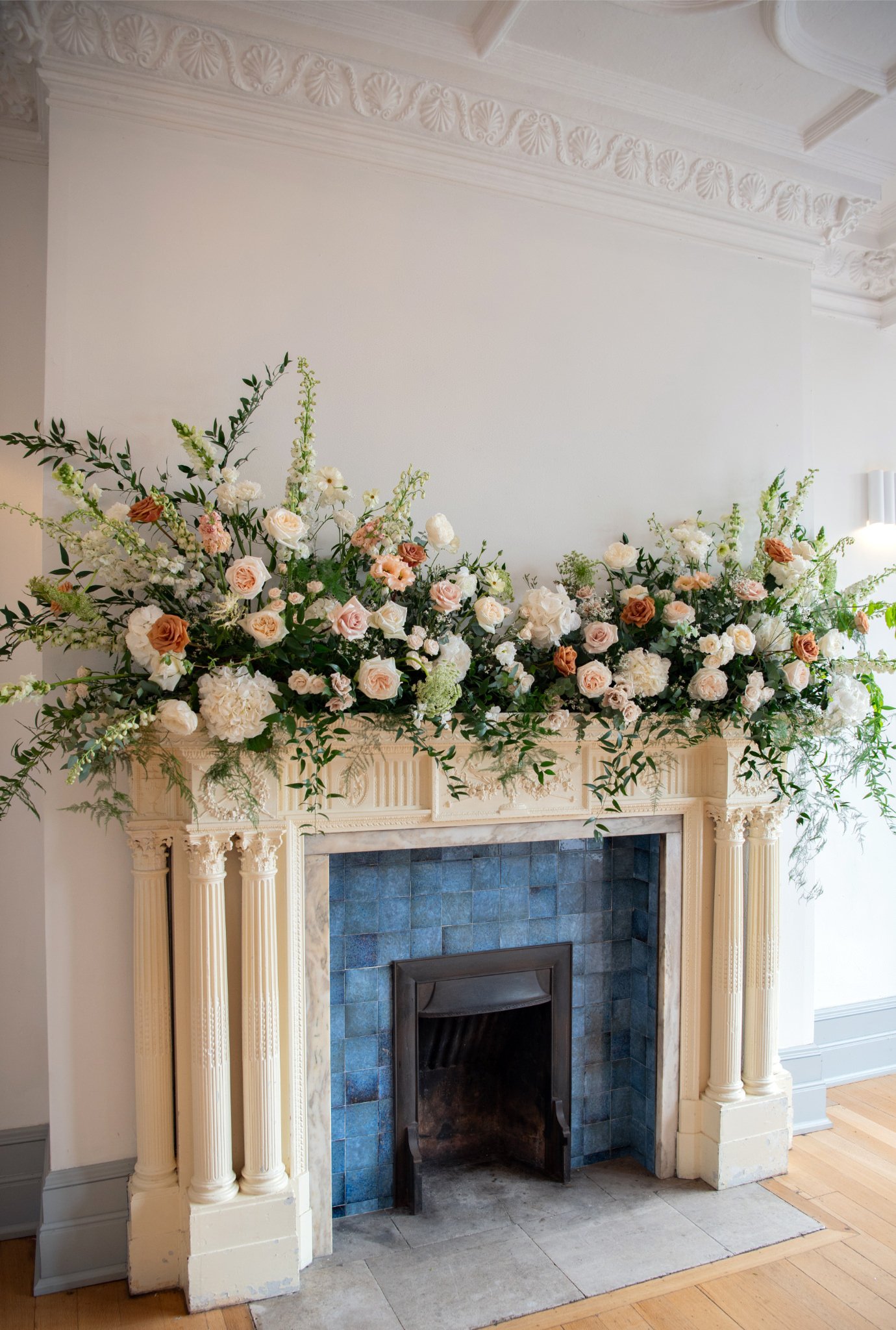 Decorative fireplace mantel adorned with a large arrangement of white and light pink flowers, including roses and hydrangeas, with green foliage, in a well-lit room.