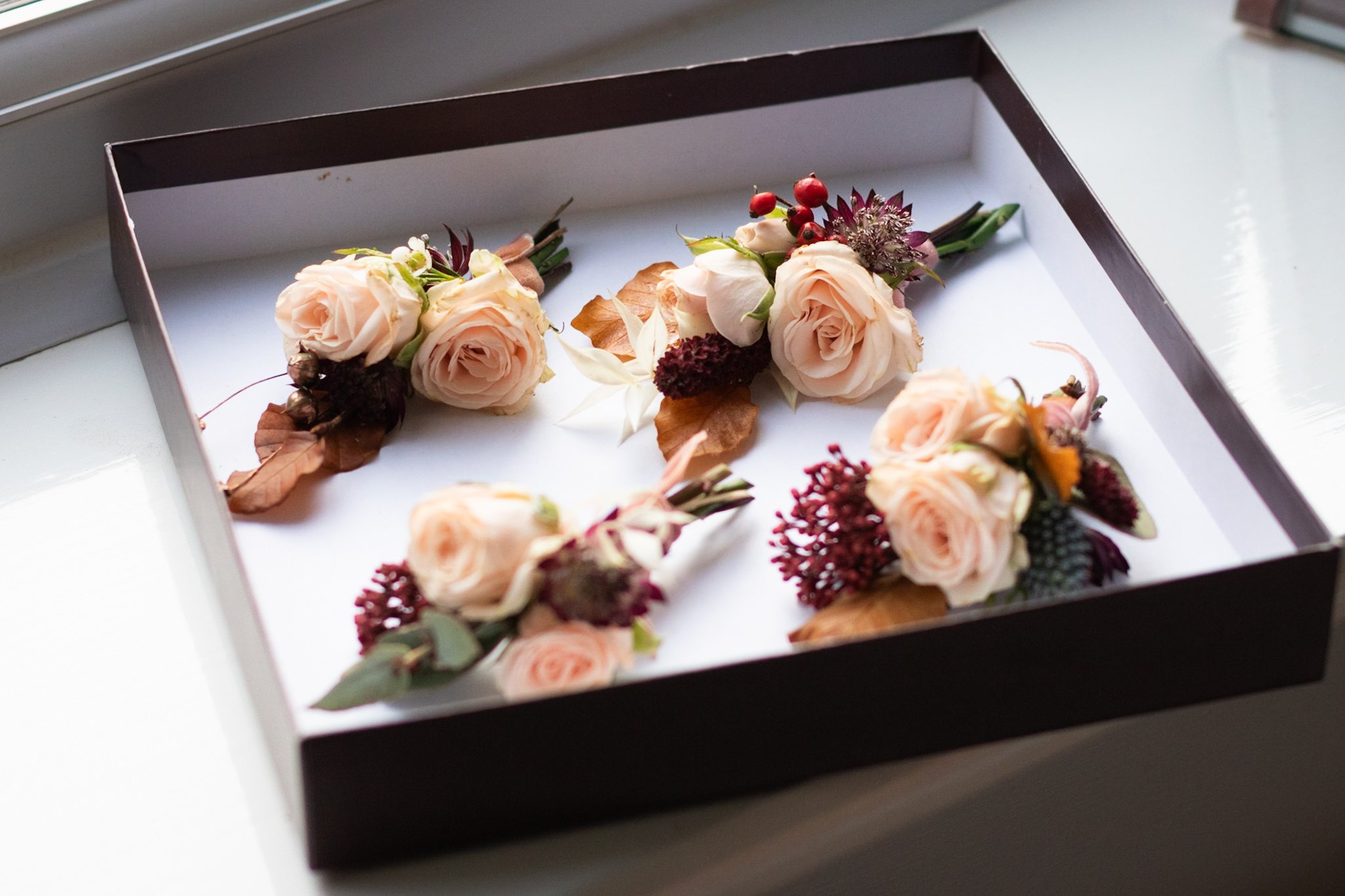 Several peach-colored rose boutonnieres with greenery and red accents laid out in a black box.