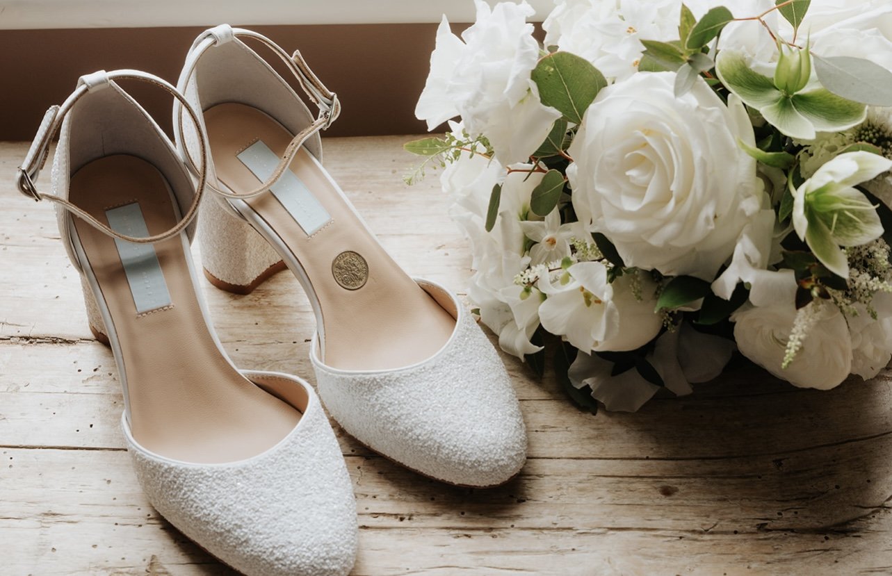 White high-heeled shoes with ankle straps next to a white floral bouquet on a wooden surface.