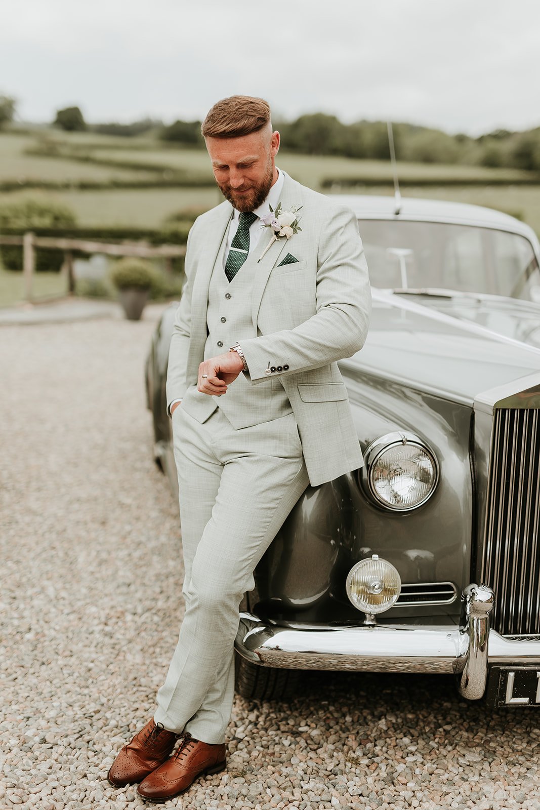 A groom in a light gray suit leaning against a vintage silver car, checking his wristwatch on a gravel driveway with a countryside background.