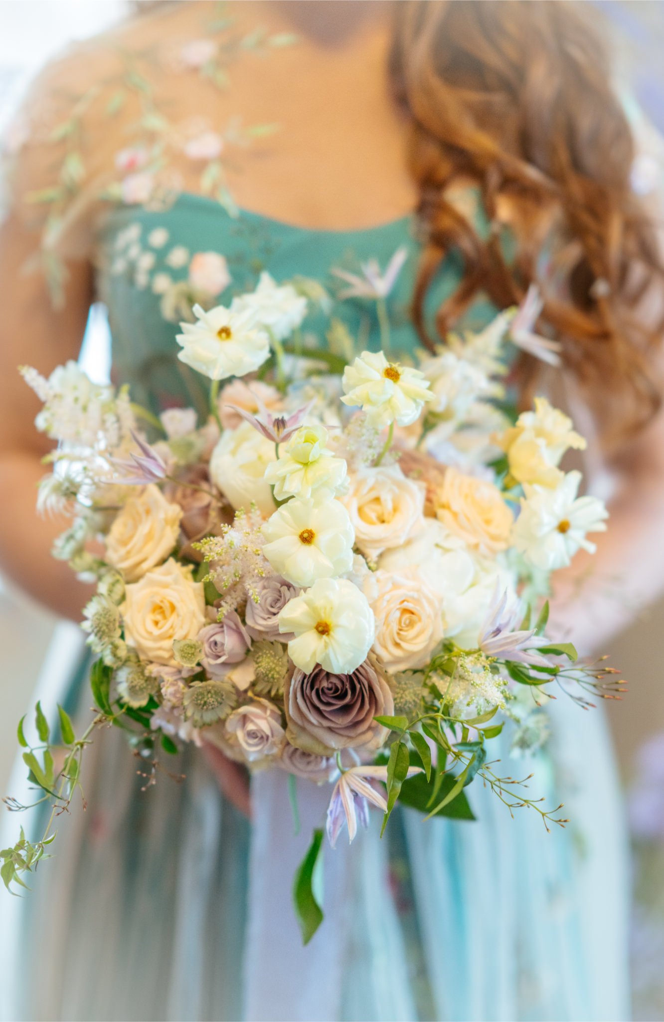 A person in a teal dress with floral embroidery is holding a large bouquet of cream, lavender, and white flowers.