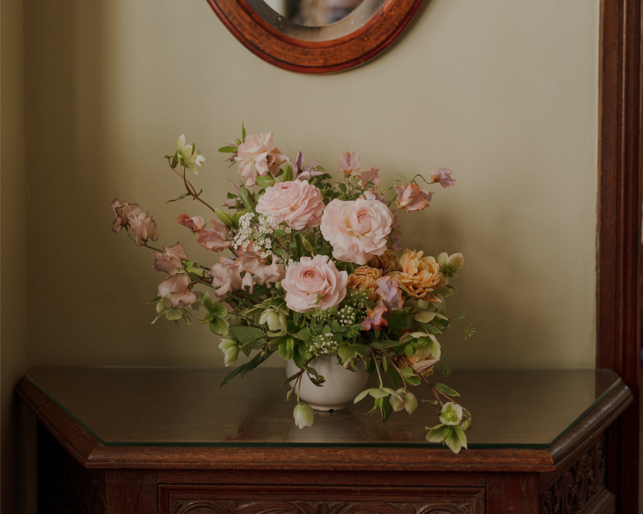 A bouquet of pink and peach flowers in a white vase on a wooden table against a green wall, with a mirror above.
