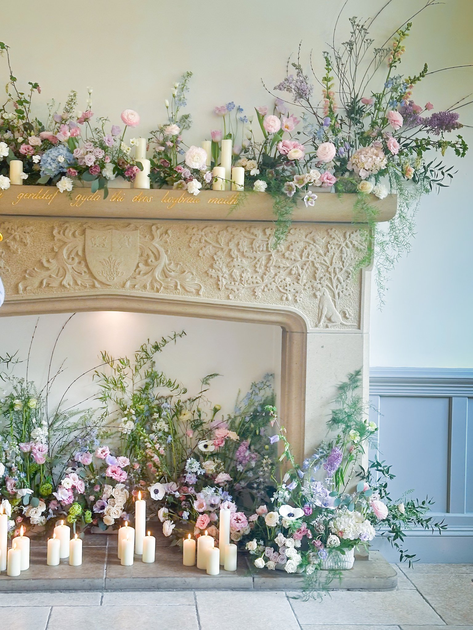 A decorated fireplace with a variety of pastel-colored flowers, white candles, and greenery on and around the mantel.
