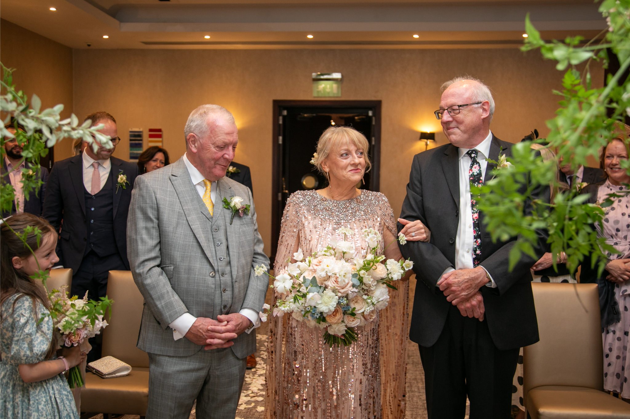 An elderly woman dressed in a sparkling, peach-colored dress holds a bouquet of white and pastel flowers while walking with two elderly men during a wedding ceremony. The man on her right is wearing a light gray plaid suit with a yellow tie and bouto