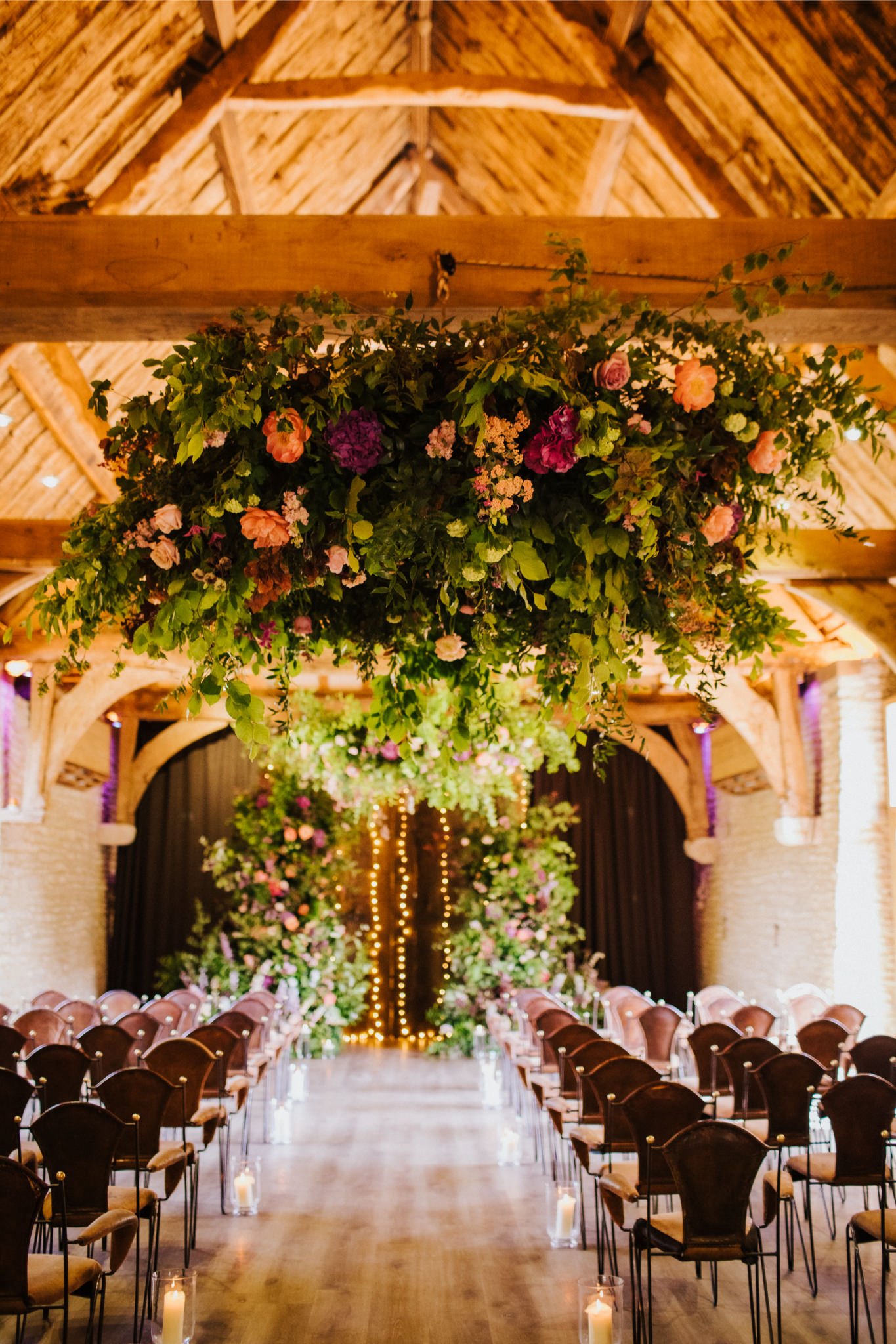 A wedding ceremony setup with rows of chairs on both sides of a wooden aisle, decorated with candles in glass holders. Overhead, large floral arrangements with pink, purple, and peach flowers hang from the wooden ceiling beams, creating a lush, roman