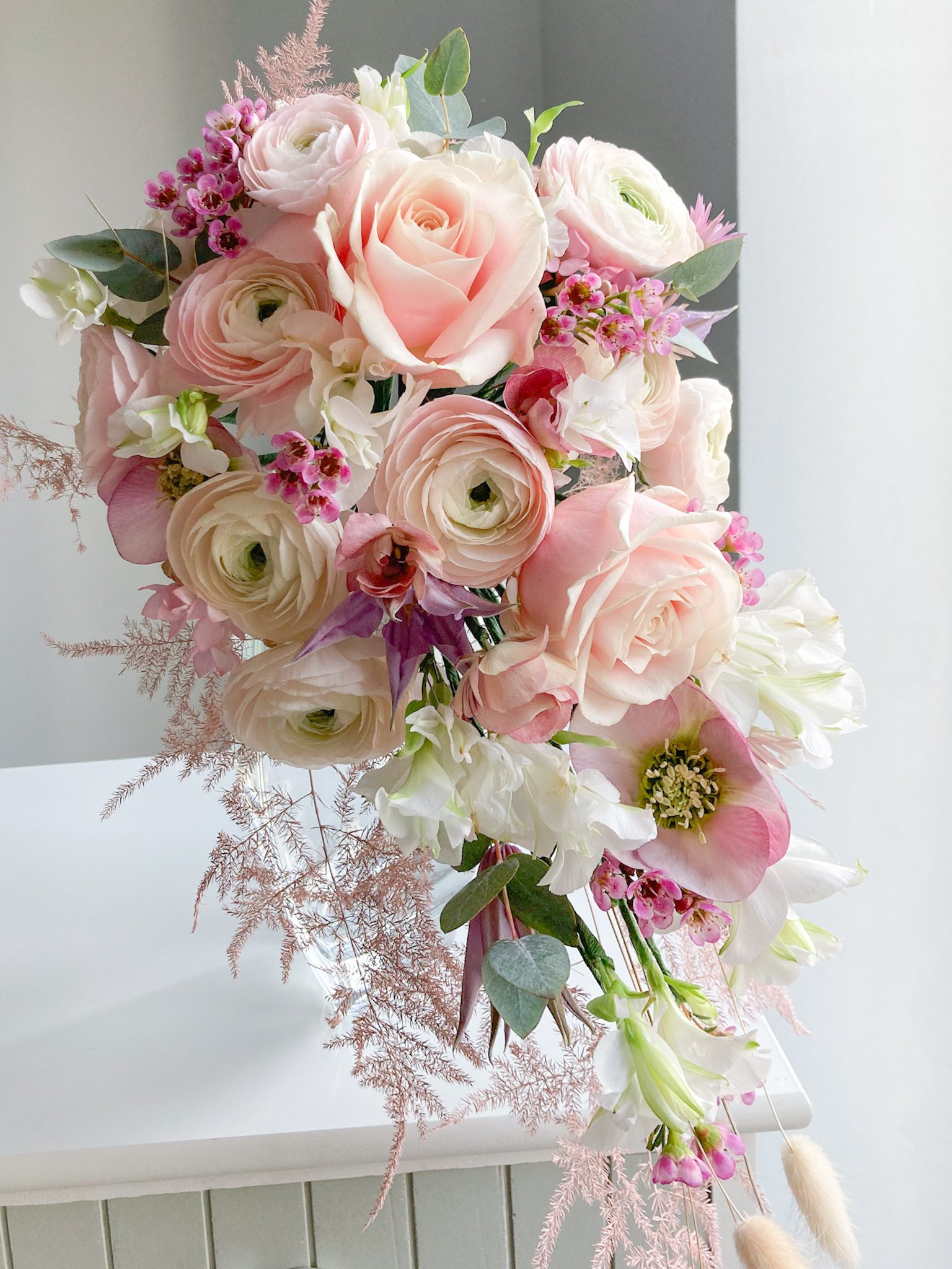A cascading bouquet of pink and white roses, ranunculus, and other flowers with greenery, sitting on a white table.