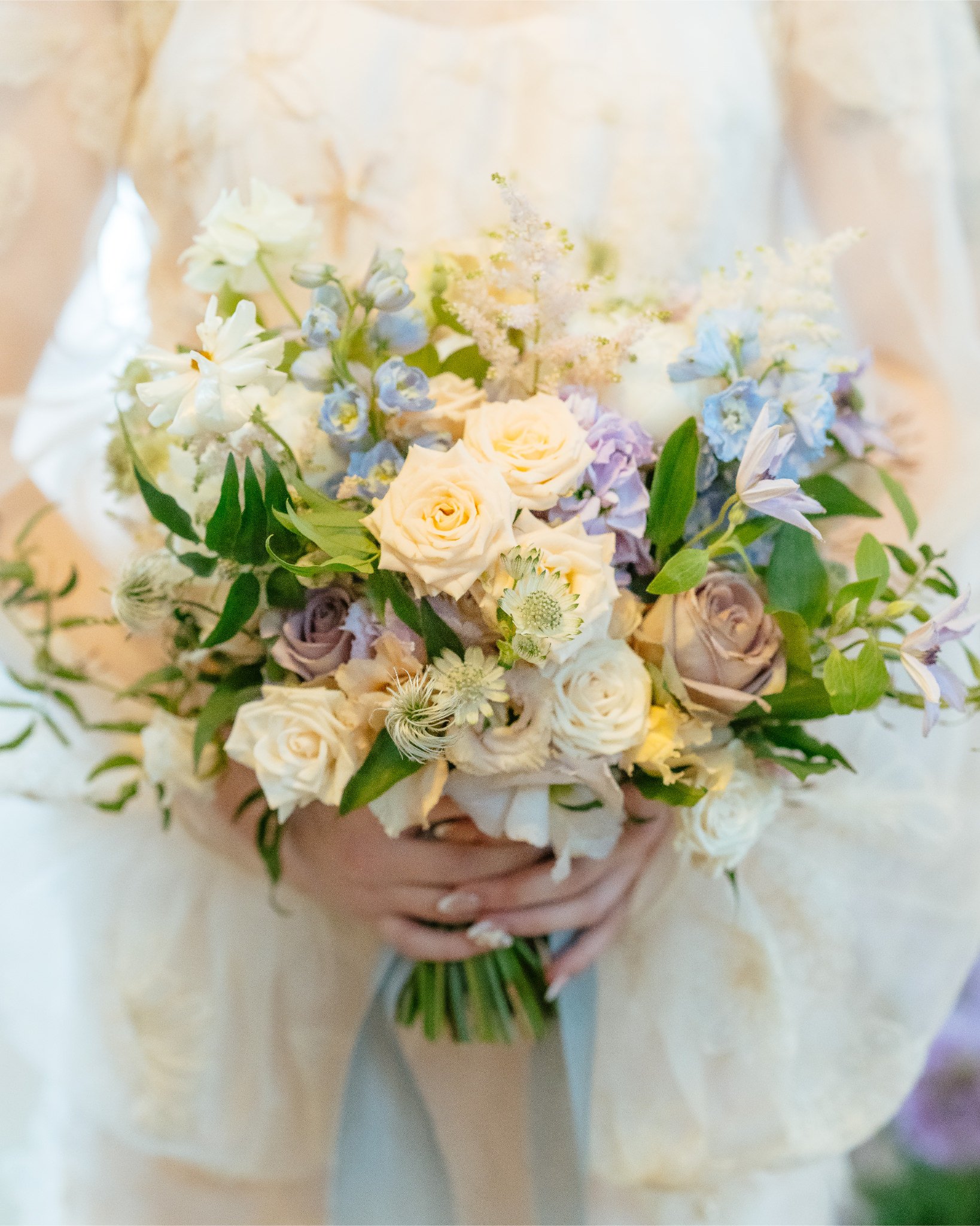 A person holding a bouquet of mixed pastel-colored flowers including roses, lisianthus, and delphiniums with green foliage, in a cream-colored dress.