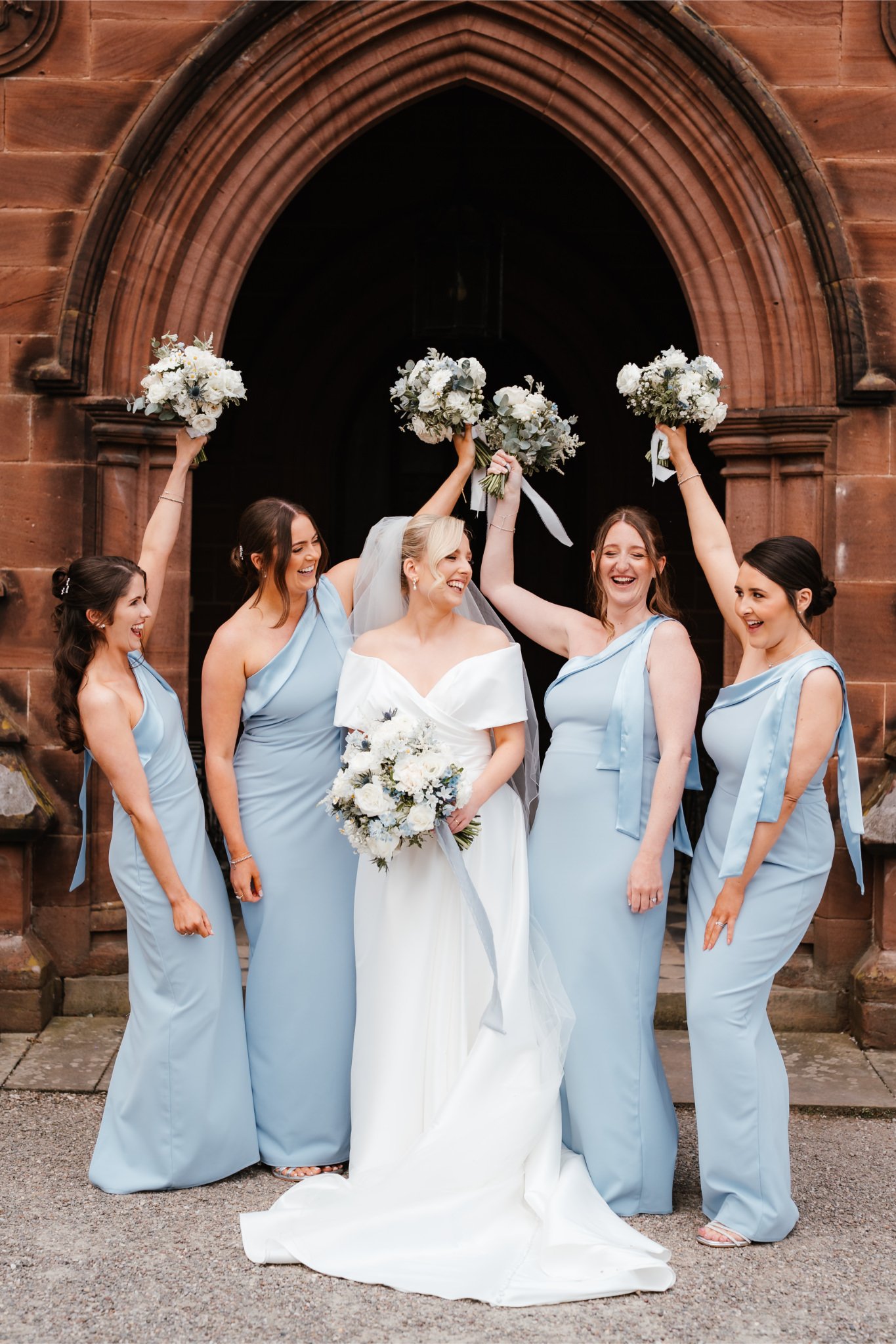 Bride and five bridesmaids celebrating in front of a church archway, with the bridesmaids raising their bouquets in the air and the bride holding her bouquet, all smiling and laughing.