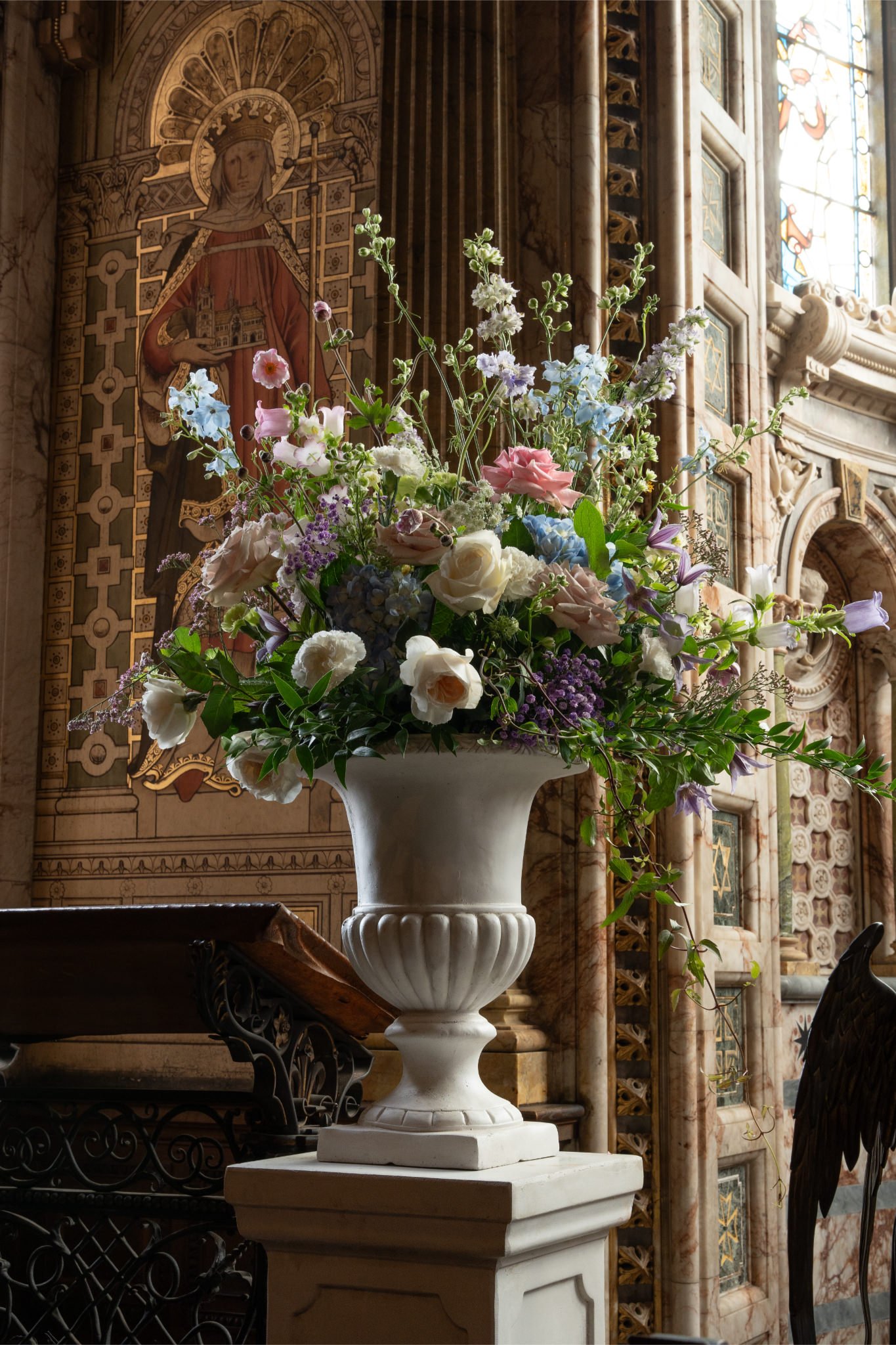 A large bouquet of assorted flowers in a white vase inside a church with ornate stained glass windows and religious artwork decorating the walls.