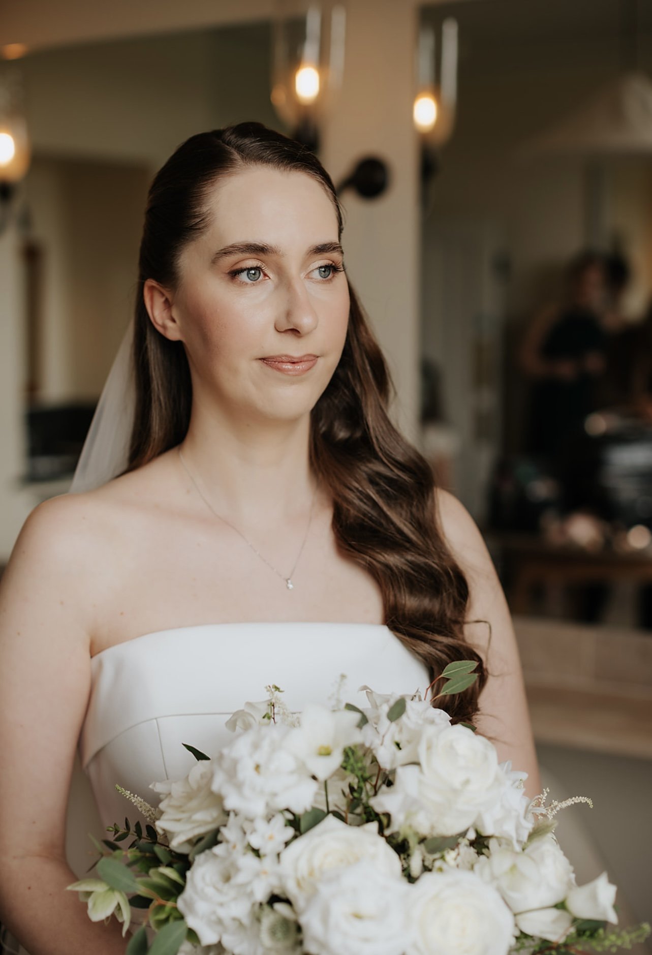 A bride with long brown hair and a strapless white wedding dress holding a bouquet of white roses and greenery, standing indoors with warm lighting.
