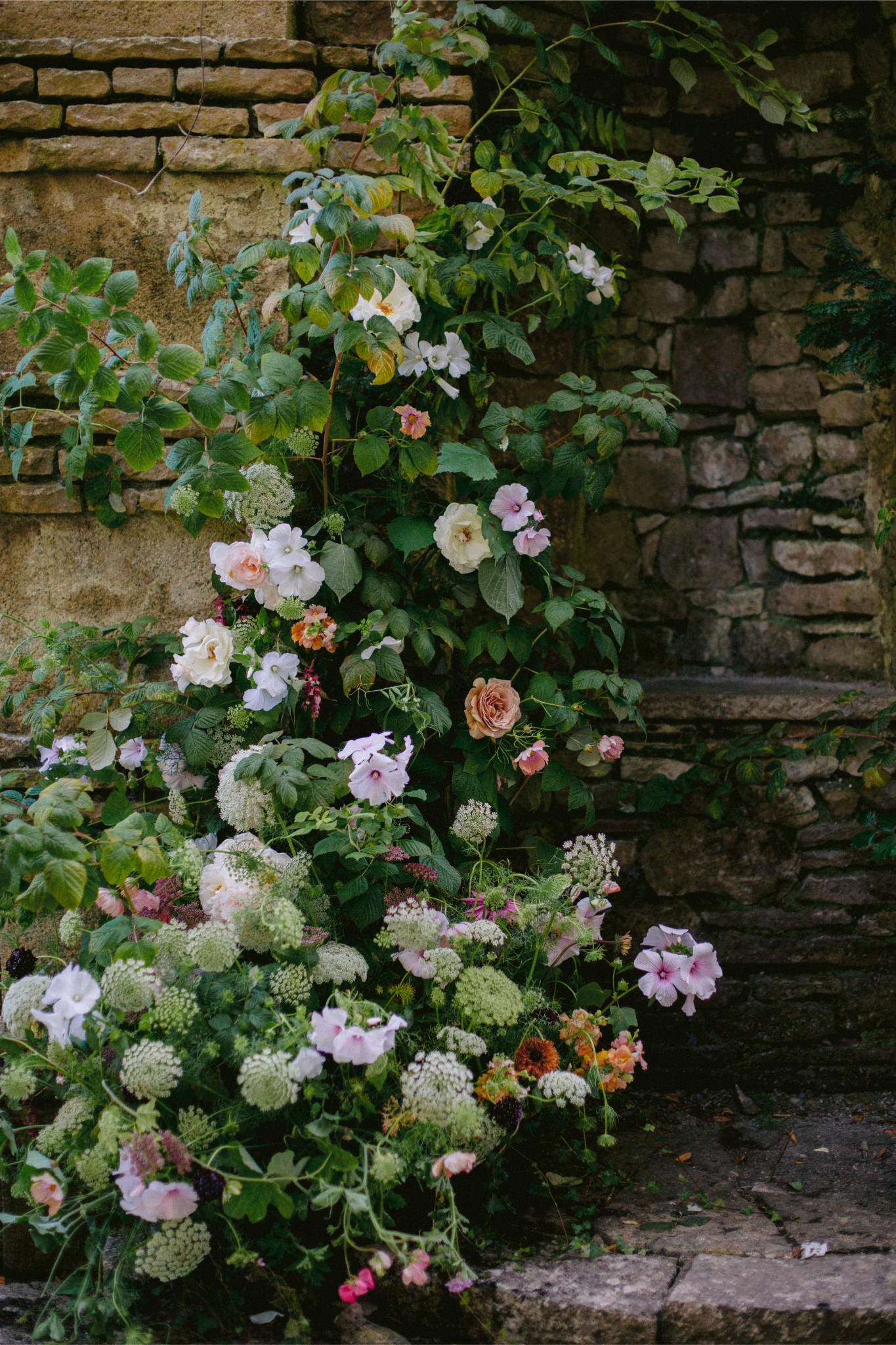 A lush garden with pink, white, and cream flowers growing against a stone and brick wall.