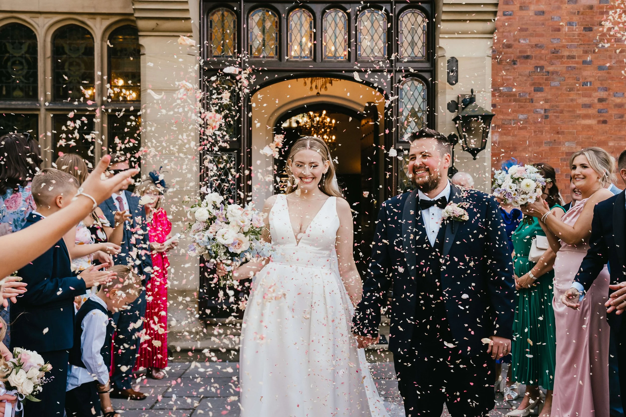 smiling happy couple with a perfect bouquet of roses from luxury weddign floristconfetti shot with oink pastel confetti and cheering family and friends