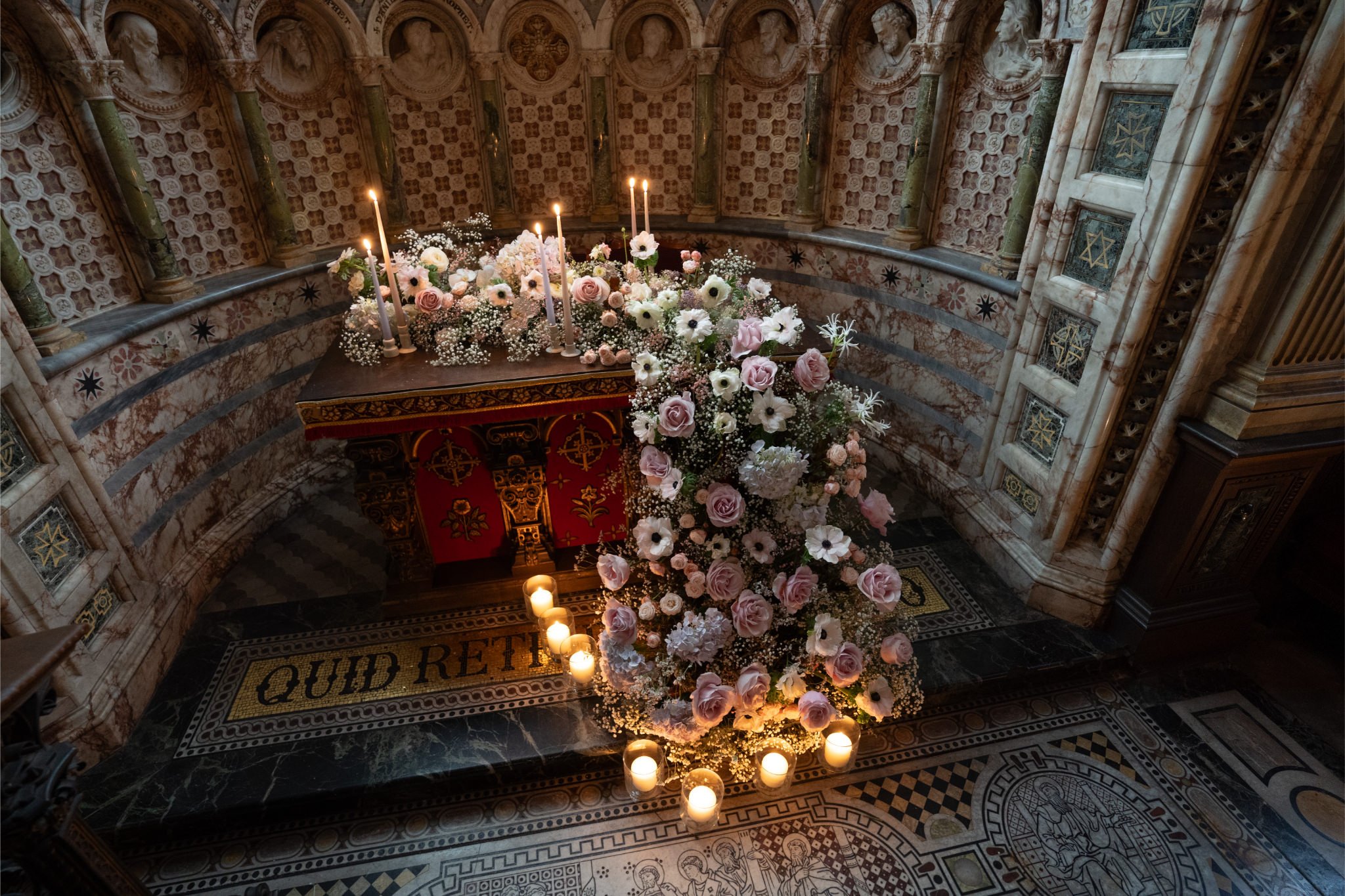 A wedding altar decorated with a large, cascading floral arrangement of pink roses, white anemones, and baby's breath, with candles surrounding it, inside a historic church with ornate patterned walls and religious artwork.