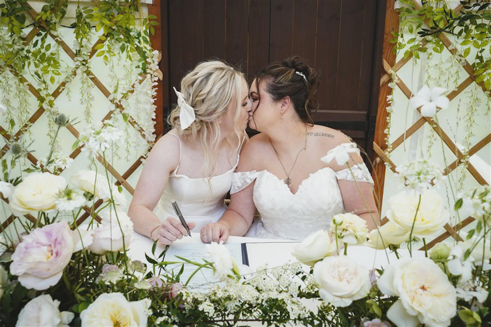 Two women in wedding dresses kiss at a wedding ceremony, surrounded by white flowers and greenery.