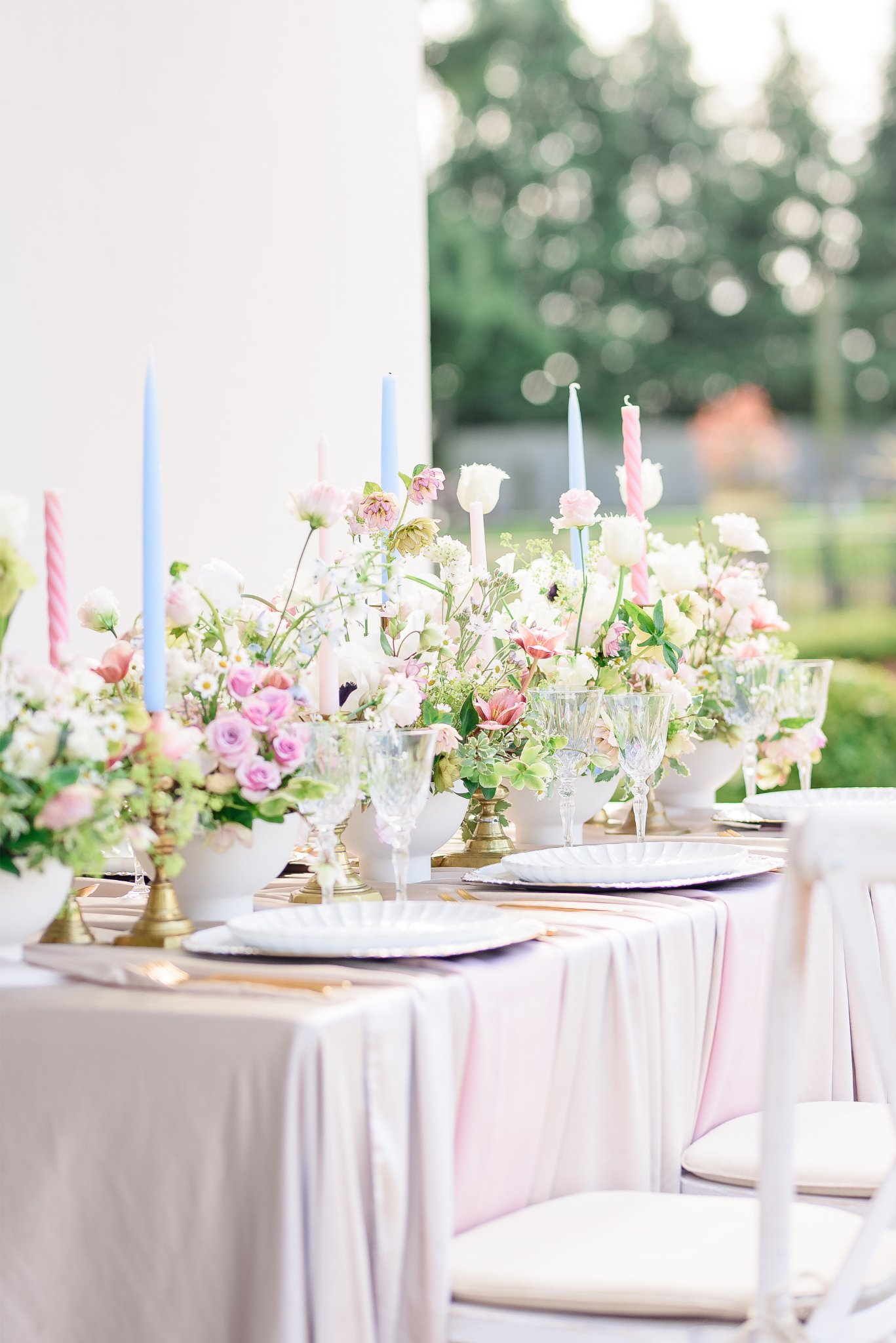 Elegant outdoor table setting with pink and white floral arrangements, colored candles, glassware, and white plates.