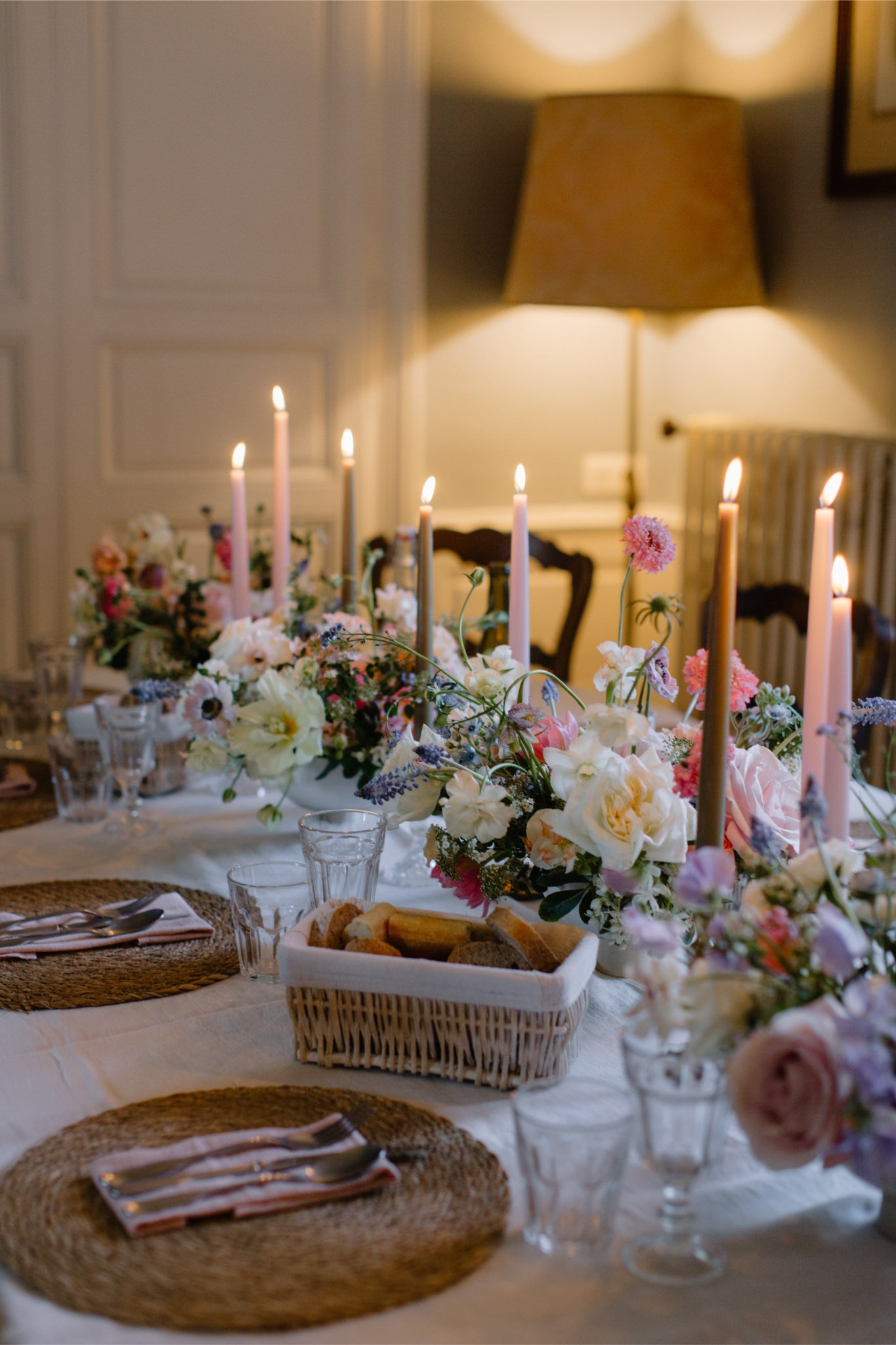 A dinner table set with floral arrangements, pink candles, glassware, silverware, and woven placemats, illuminated by soft lighting in a cozy room.