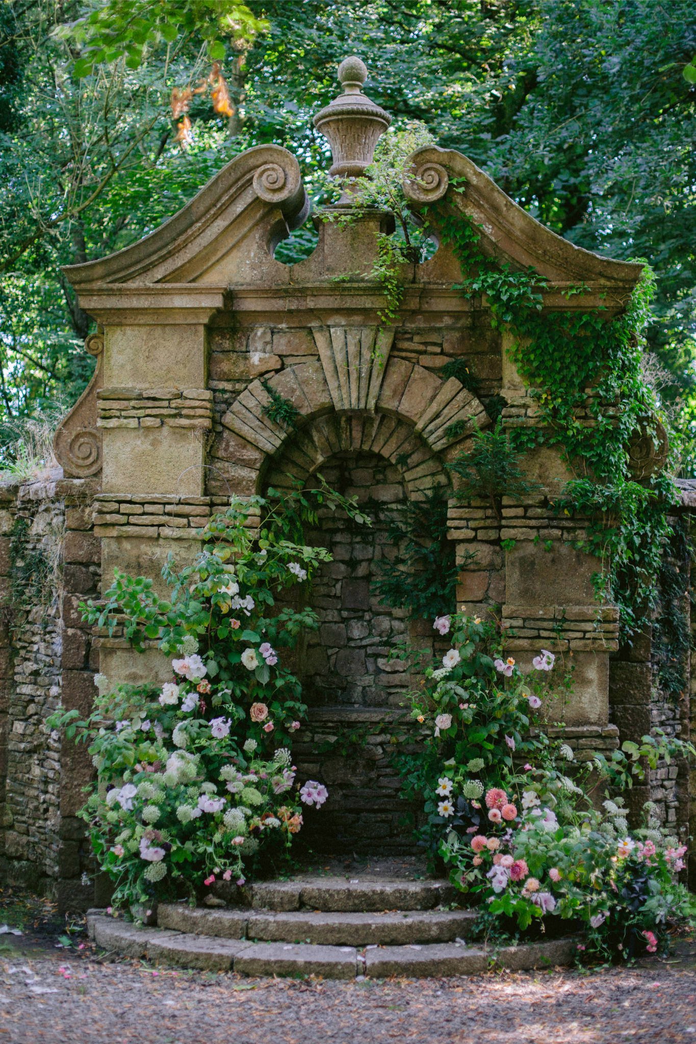 An ornate stone structure with a rounded arch at its center, decorated with greenery and flowers, surrounded by lush green trees.