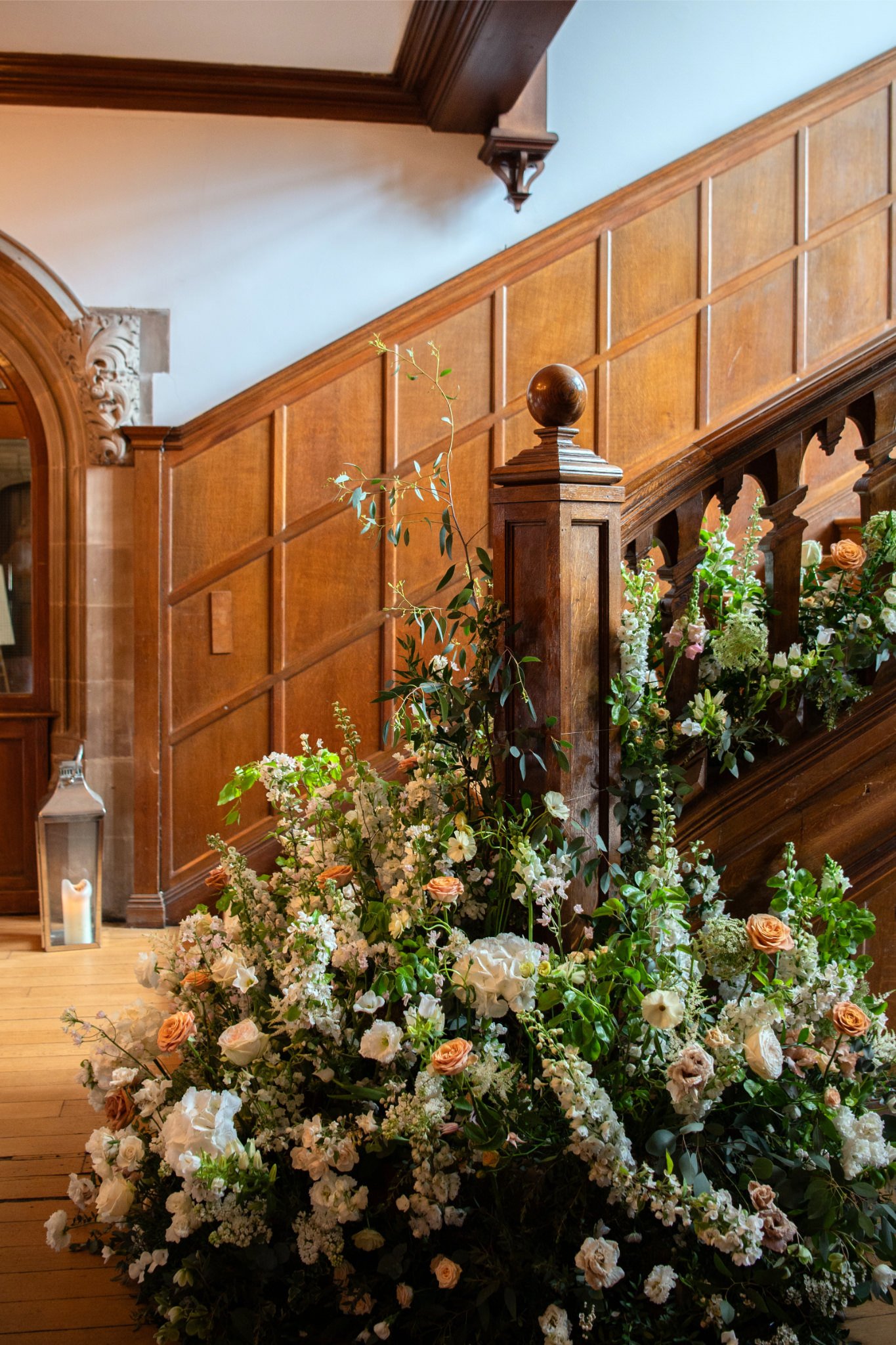 A large floral arrangement with white and peach-colored flowers decorates the base of a wooden staircase in a historic building.