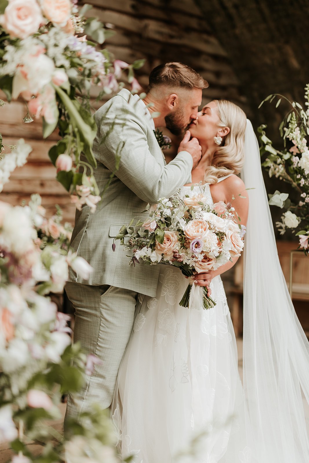 A couple sharing a kiss on their wedding day, surrounded by floral decorations, with the bride holding a large bouquet of pink and white flowers.