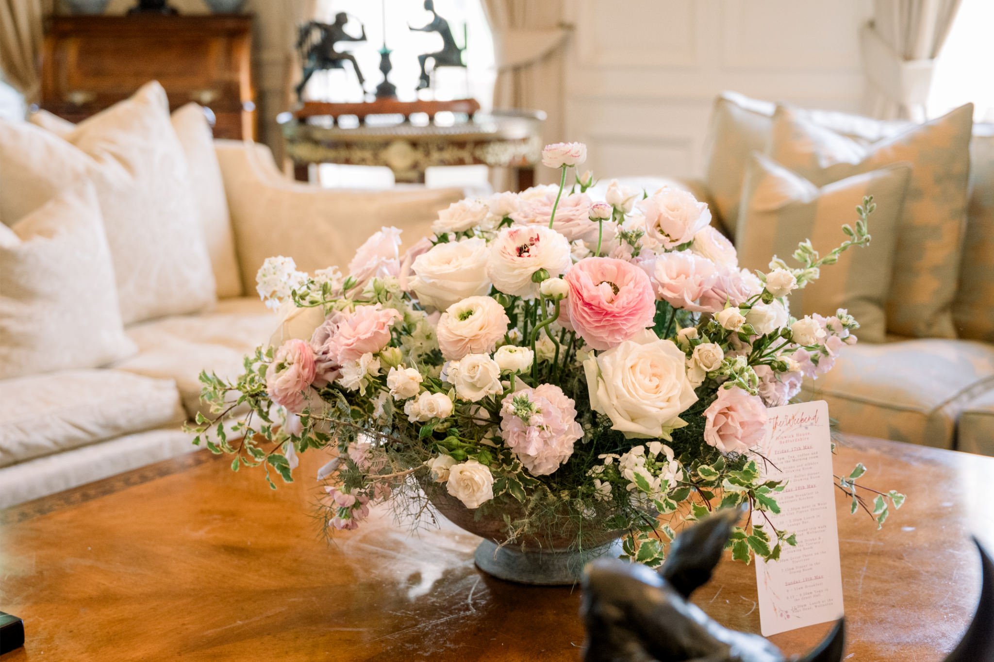 A floral centerpiece with pink and white roses and other flowers on a wooden table in a cozy, elegant living room with beige sofas and decorated walls.