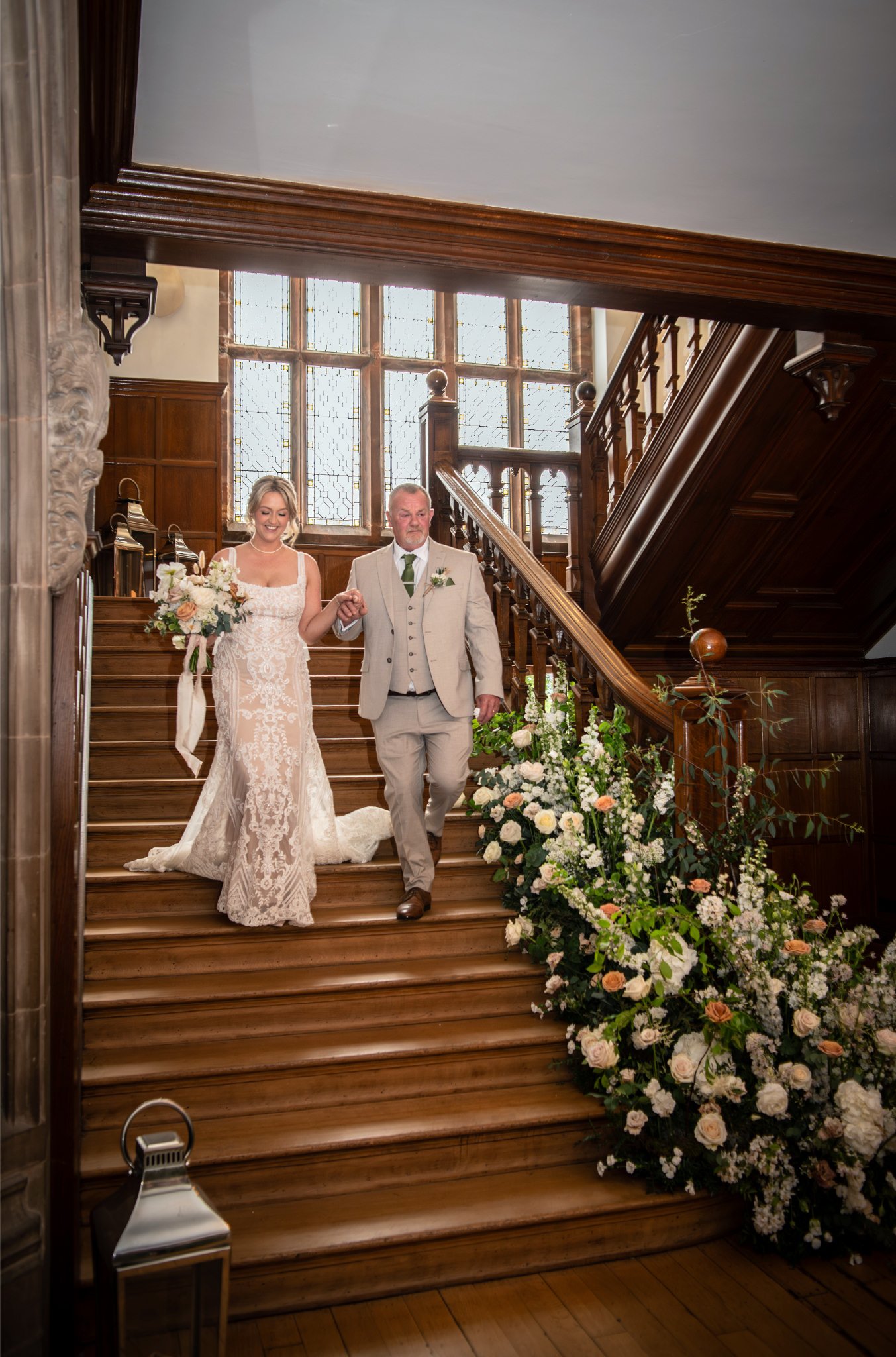 Bride and man walking down a grand wooden staircase inside a historic building, with large stained glass windows behind them, surrounded by floral arrangements.