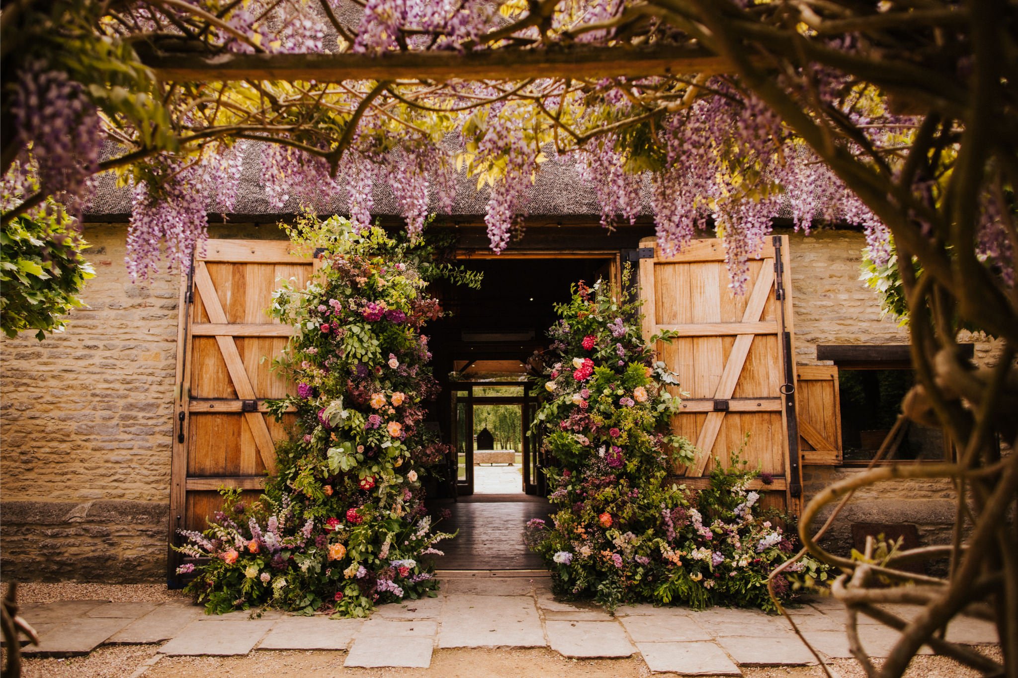 A rustic barn door adorned with vibrant floral arrangements, surrounded by purple wisteria flowers hanging overhead.