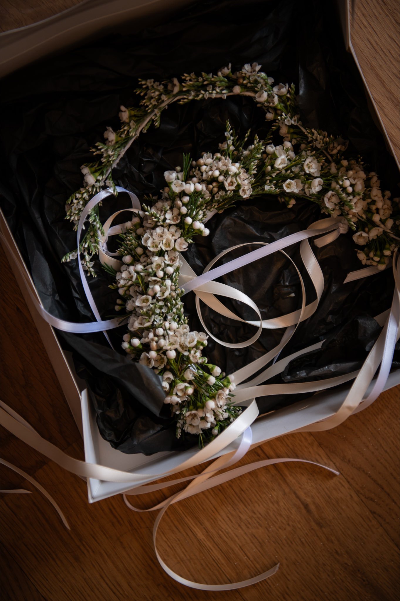 A white box containing a flower crown made of small white flowers and greenery, with white ribbons attached, placed on a wooden surface.