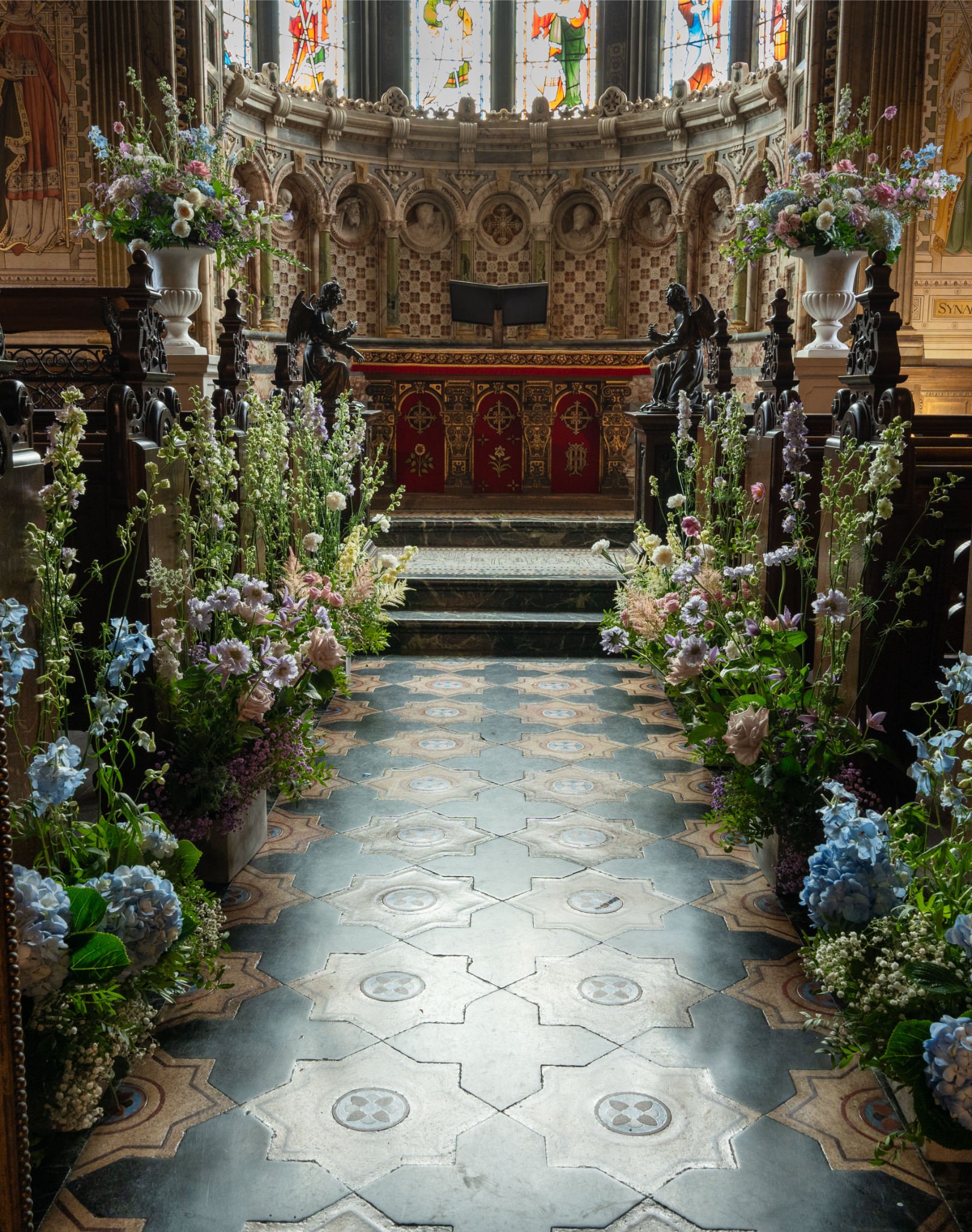 Inside a church with floral arrangements flanking the aisle, leading to an altar with statues and stained glass windows.