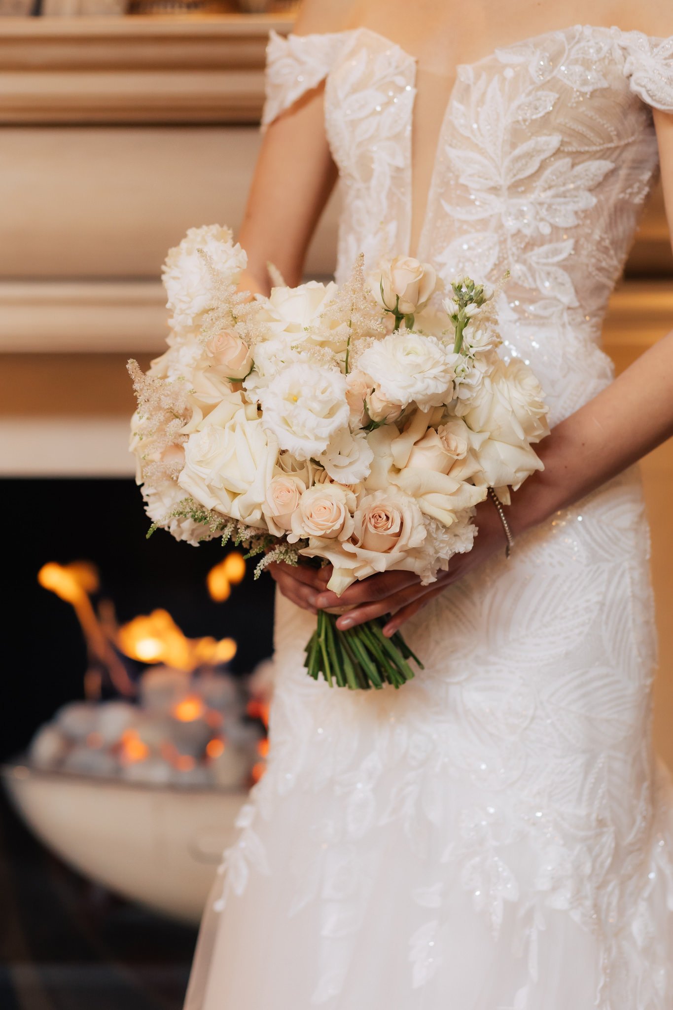 Bride in a detailed white wedding dress holding a bouquet of white and pale pink roses and flowers, standing in front of a fireplace.
