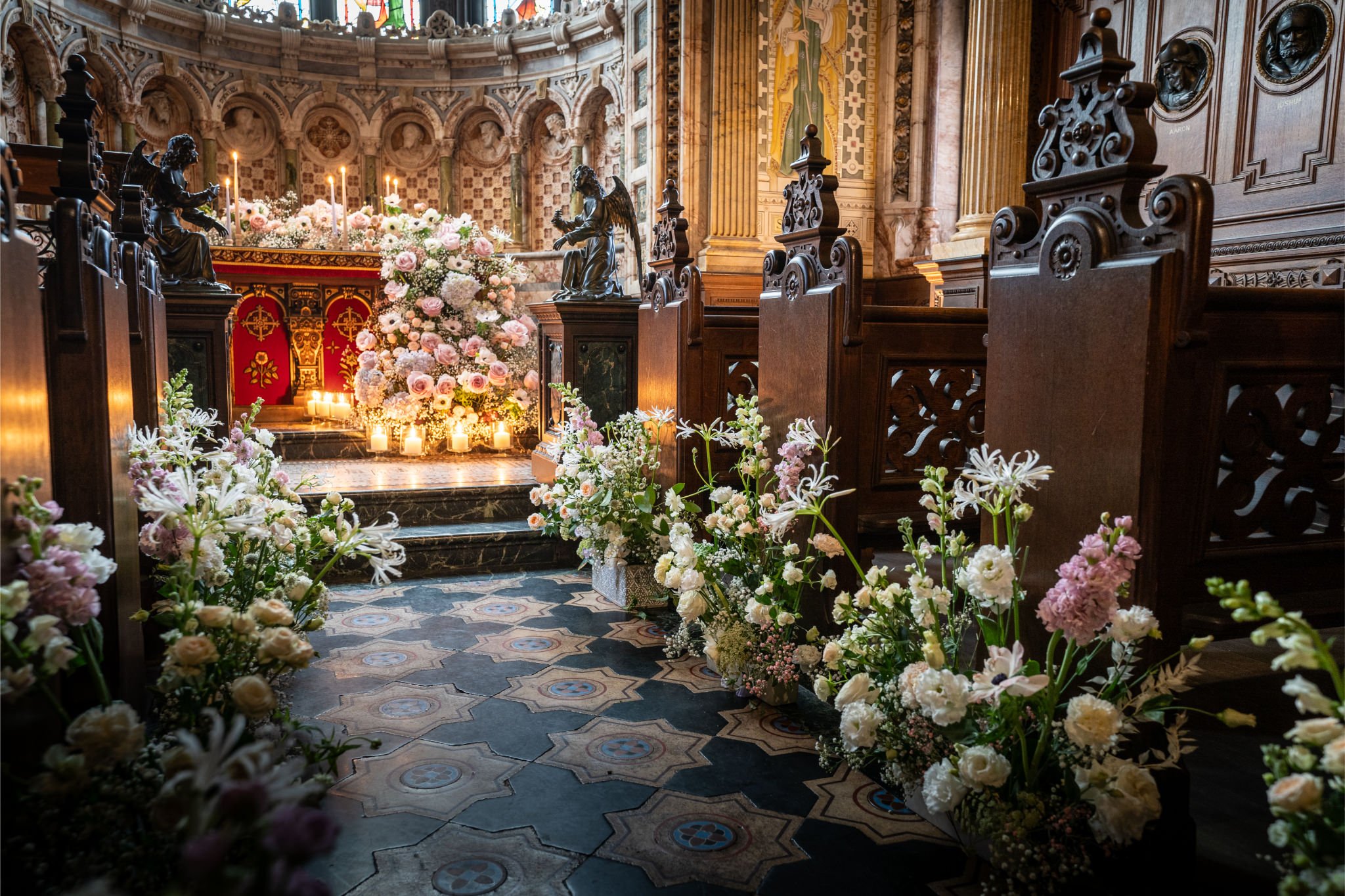 A church altar decorated with pink and white flowers, candles, and religious statues, with wooden pews and ornate architectural details.