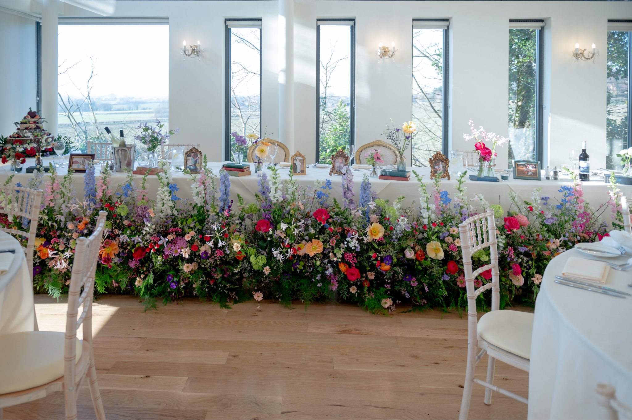 A banquet table decorated with colorful flowers, picture frames, and champagne bottles in a bright room with large windows and wood floors.
