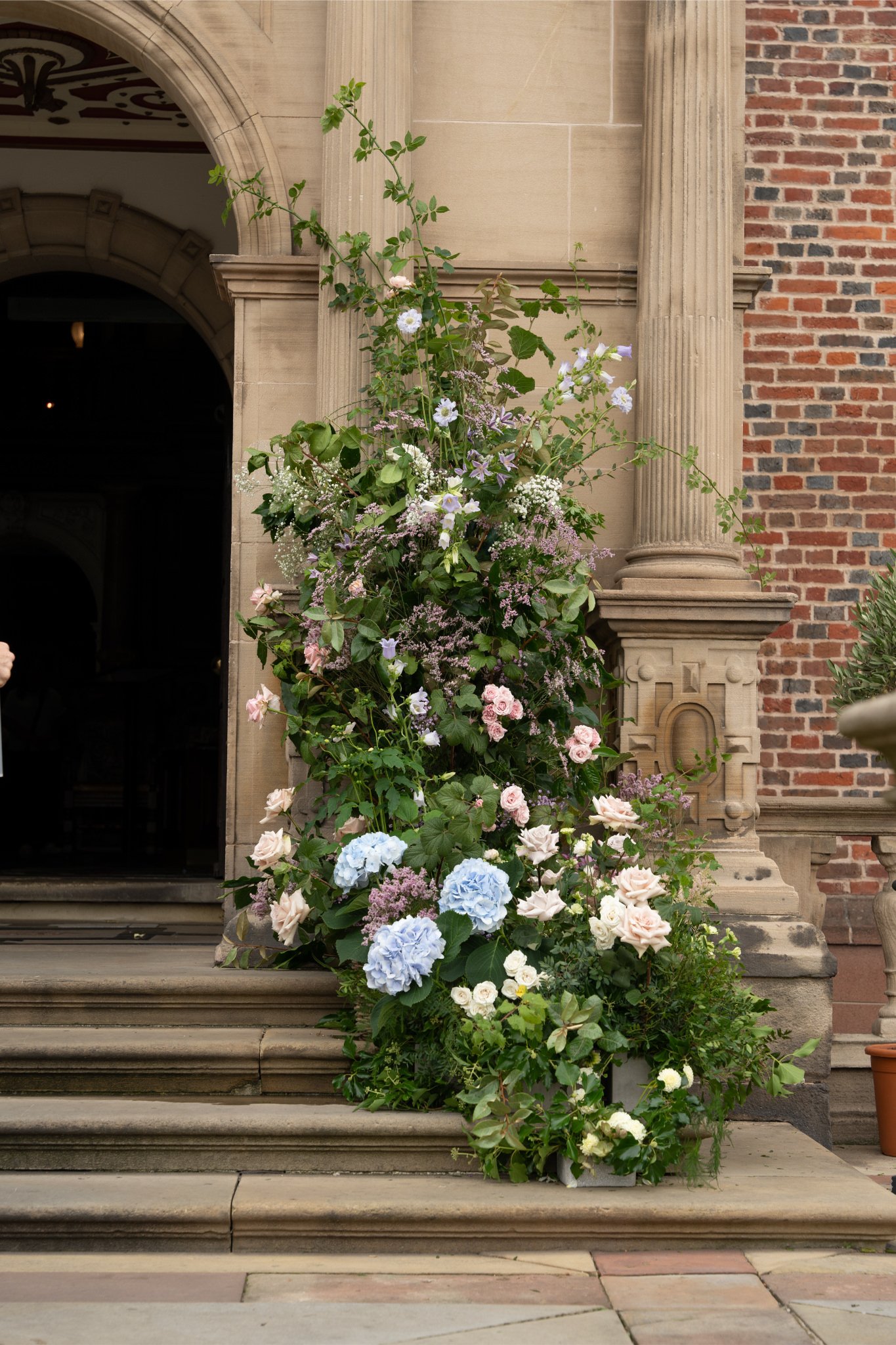 A large floral arrangement with pink, white, purple, and blue flowers on stone steps outside a historic building with brick and stone architecture.