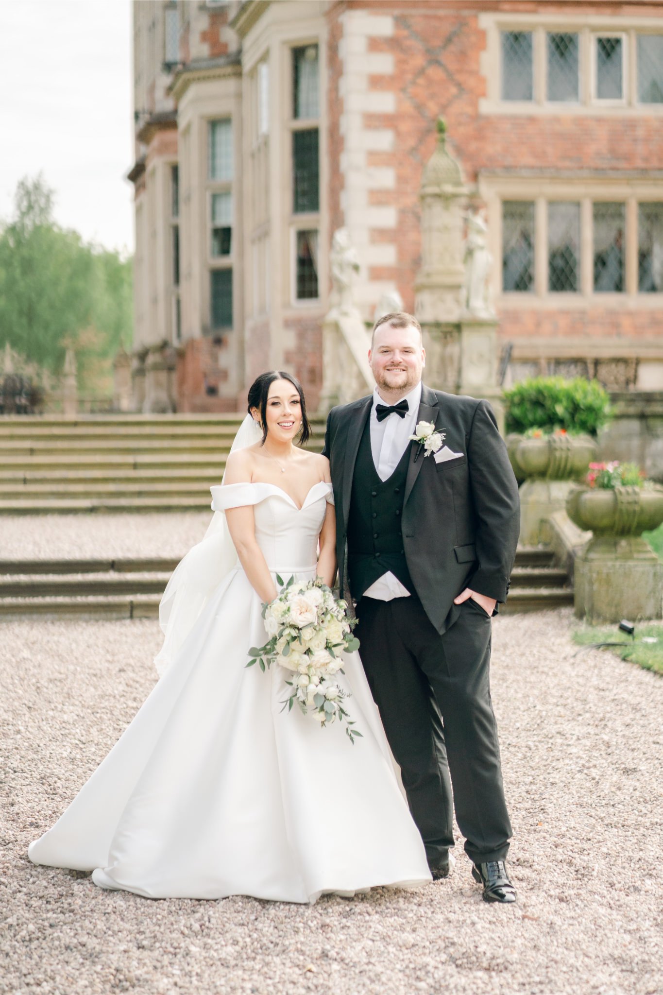Bride and groom standing together outdoors on their wedding day, smiling, in front of a historic brick building with steps, greenery, and large stone planters.