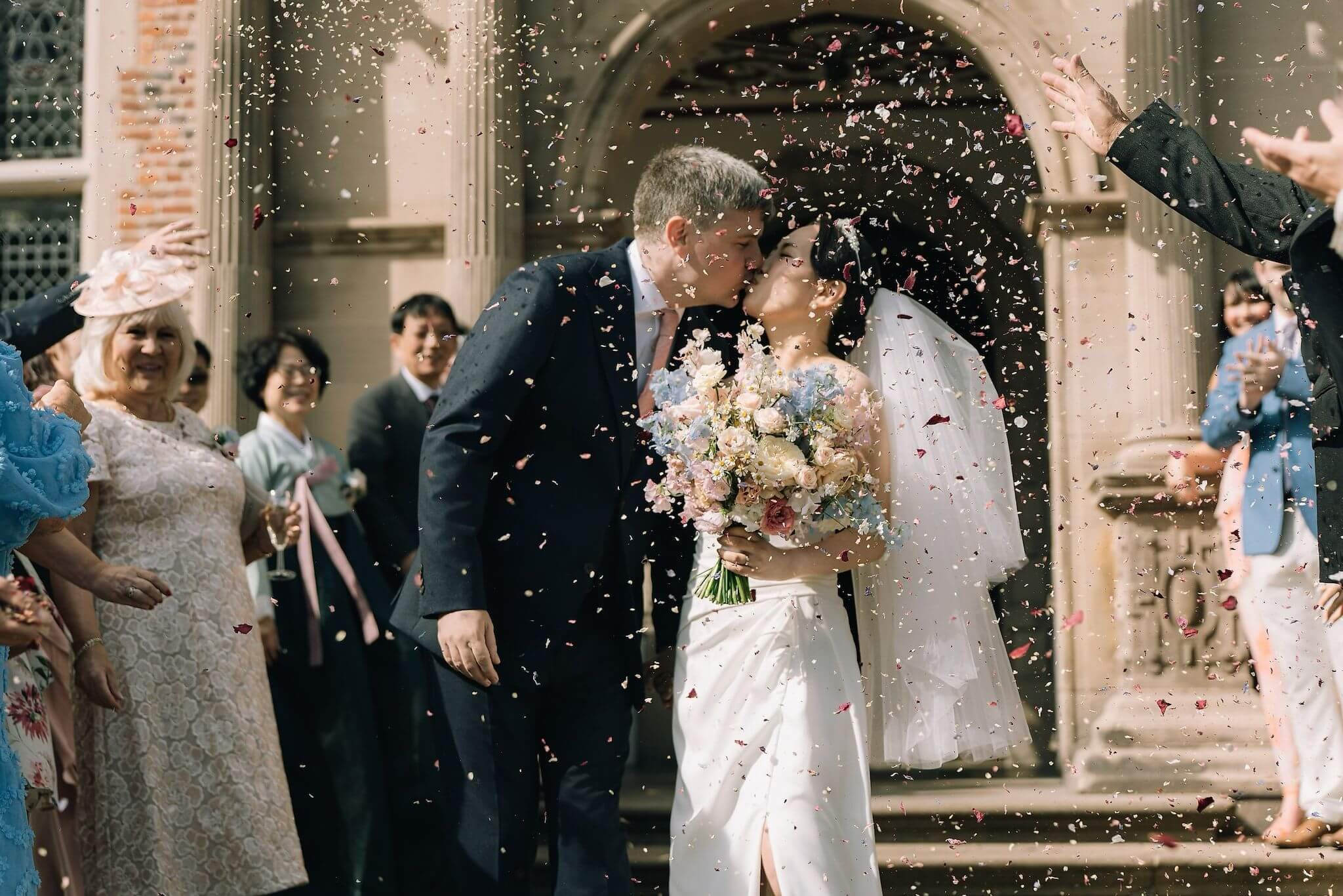 A newlywed couple sharing a kiss under a flower archway as guests celebrate around them, throwing confetti at their wedding outside a church.